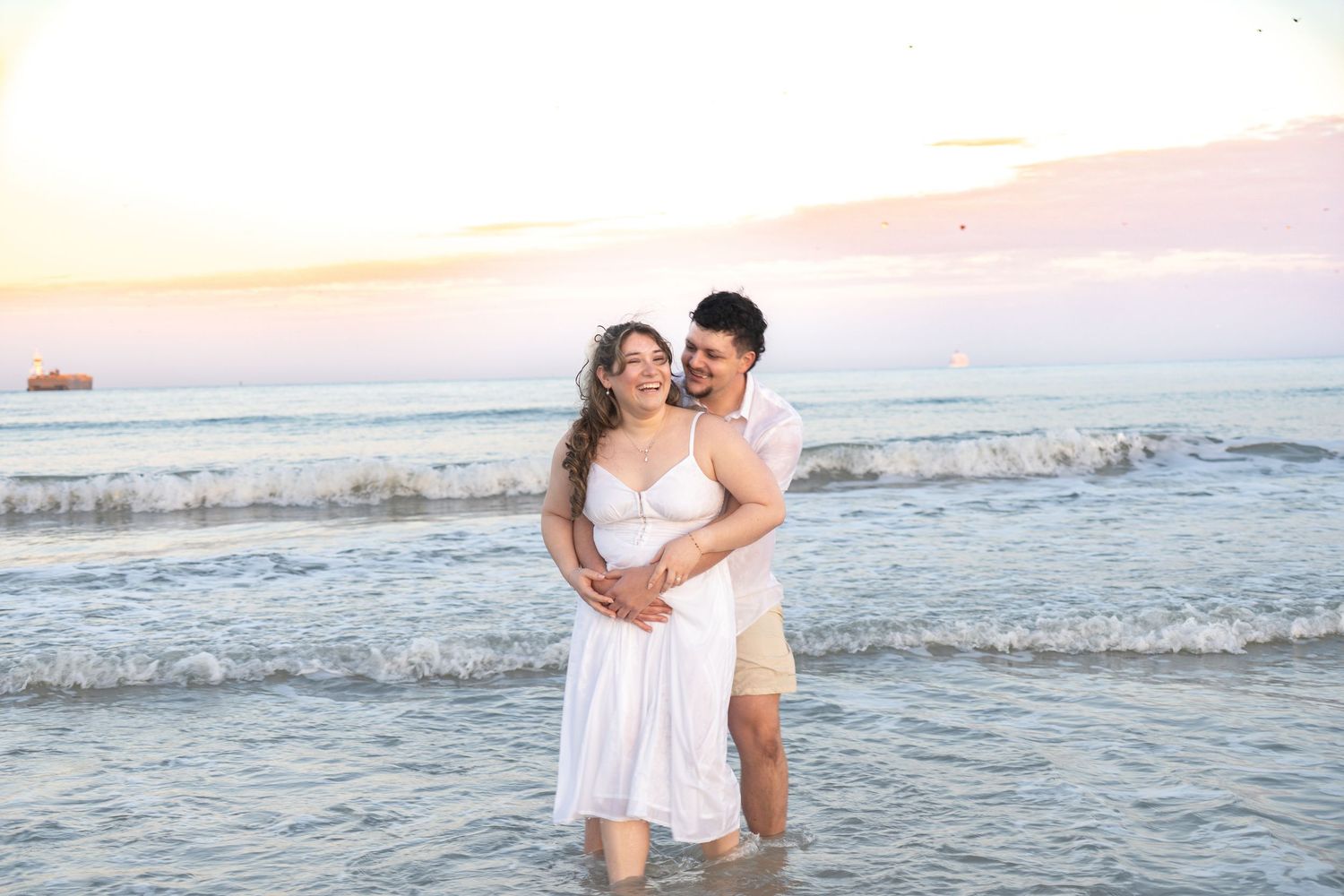 Couple embracing and smiling on beach at sunset, woman in white dress, man in pink shirt and tan shorts.