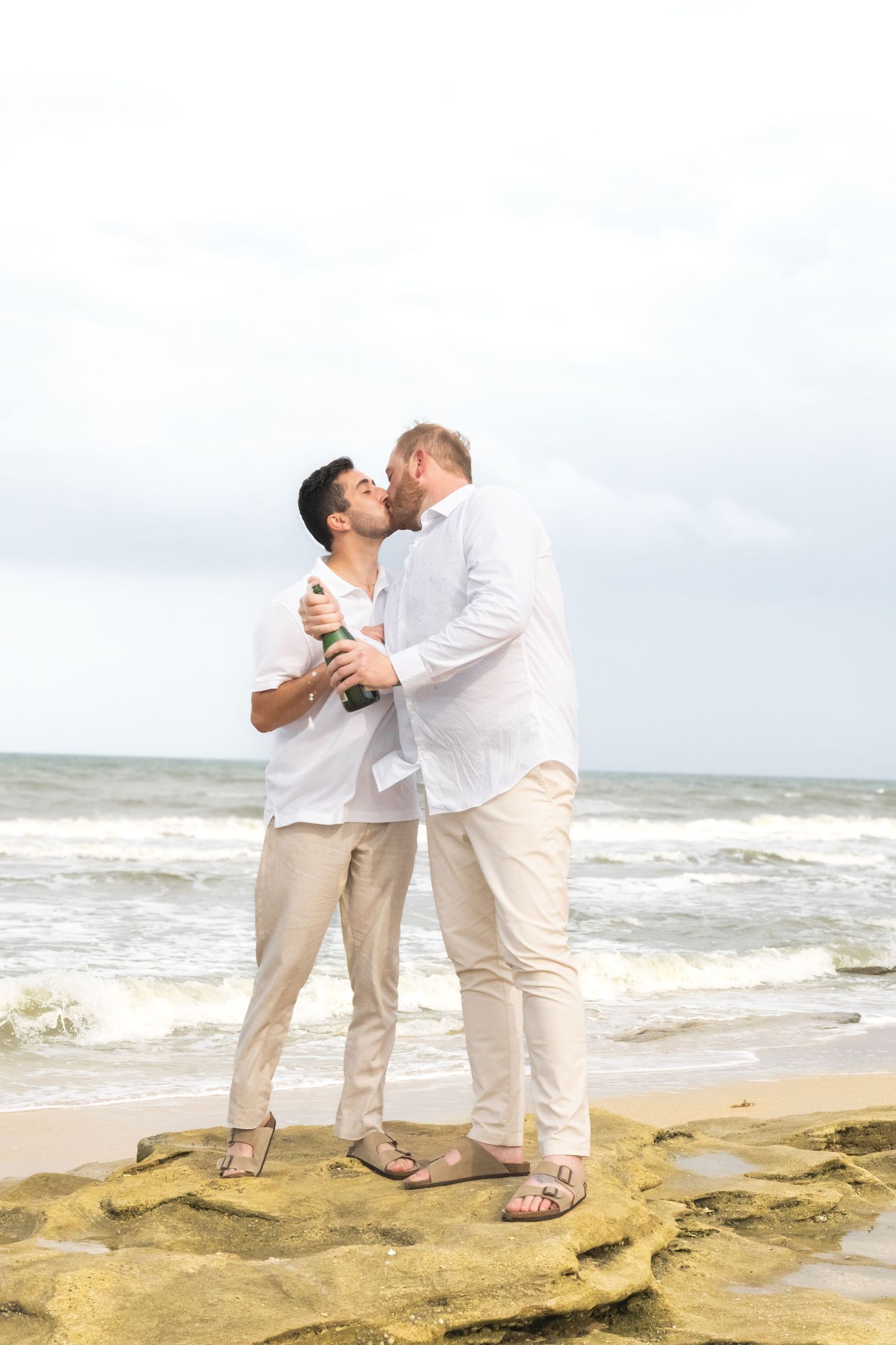 Two men kissing on a rocky beach while holding a champagne bottle, dressed in white shirts and beige pants.