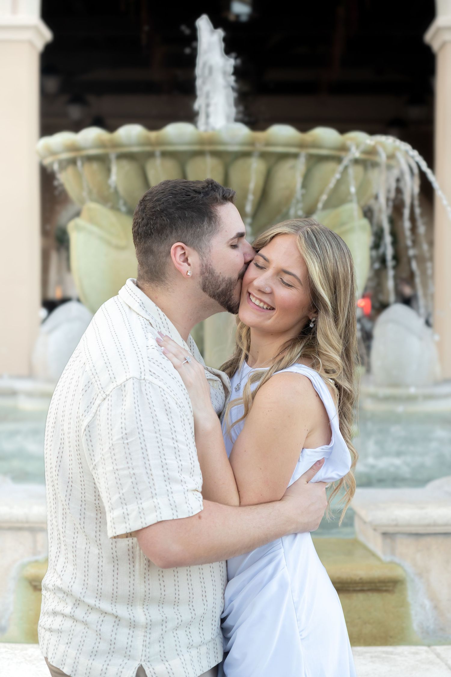 Couple embracing and kissing near a stone fountain, woman smiling joyfully in white dress.