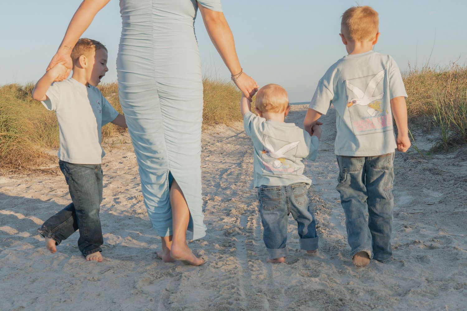 Pregnant mother in blue dress walking on sandy beach holding hands with three young children.
