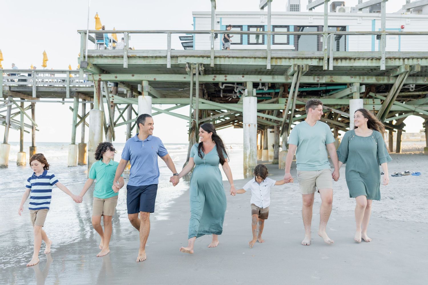 Extended family holding hands and walking along the beach shore near a pier.