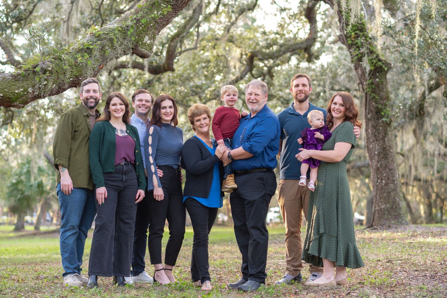 Large family group portrait outdoors under moss-covered oak trees on a sunny day in a lush green park.