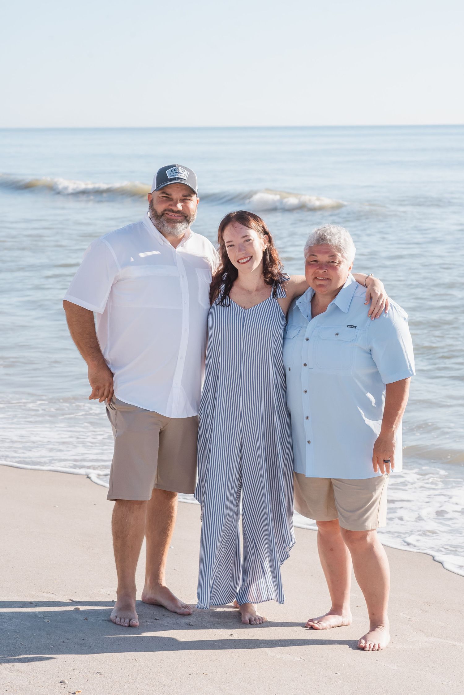 Three smiling people pose together on a sunny beach with ocean waves in the background.