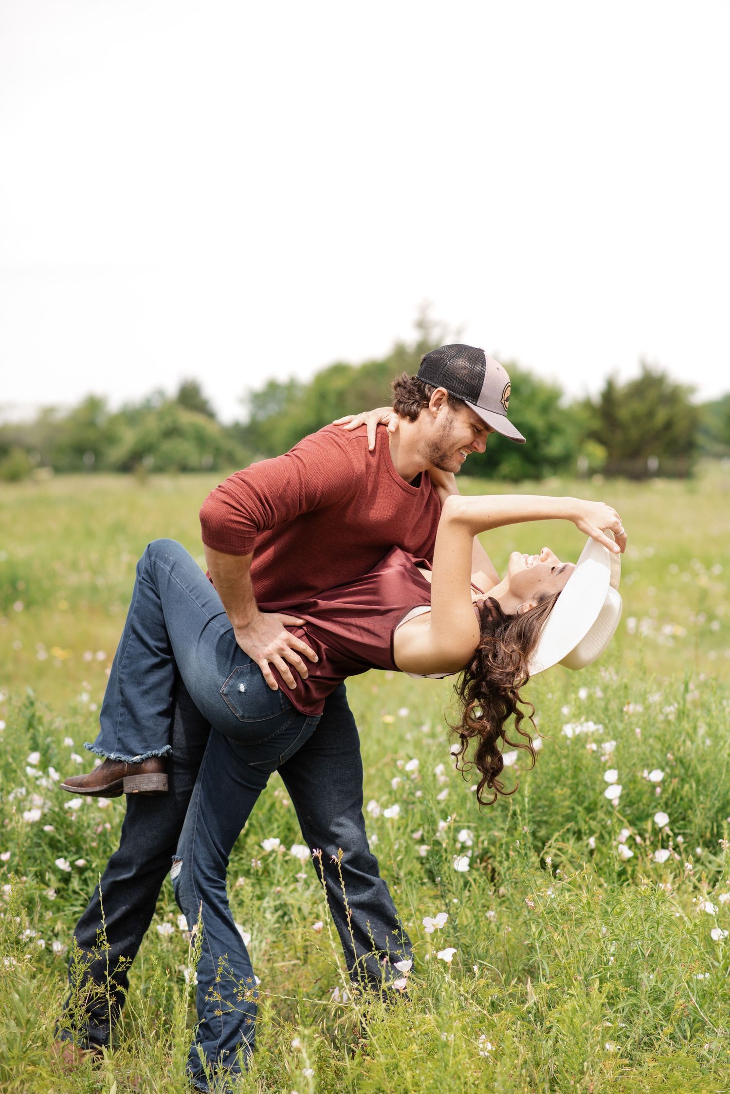 A couple dances romantically in a wildflower field, the man dipping the woman in a white hat outdoors.