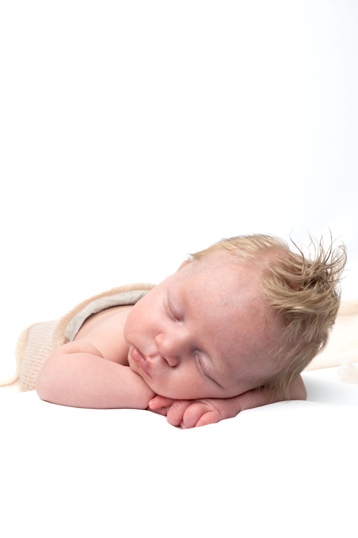 A peaceful newborn baby with blonde hair sleeping on a white background, resting cheeks on tiny hands.