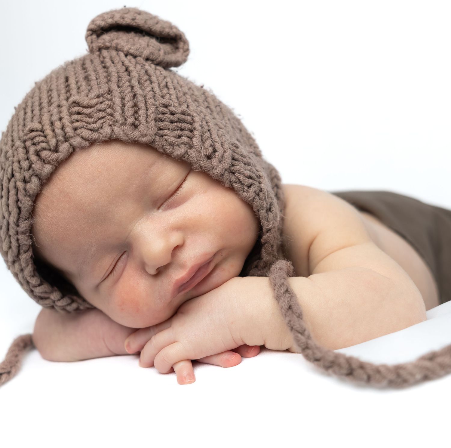 Sleeping newborn baby wearing a cozy brown knitted bonnet hat, resting peacefully on hands against white background.