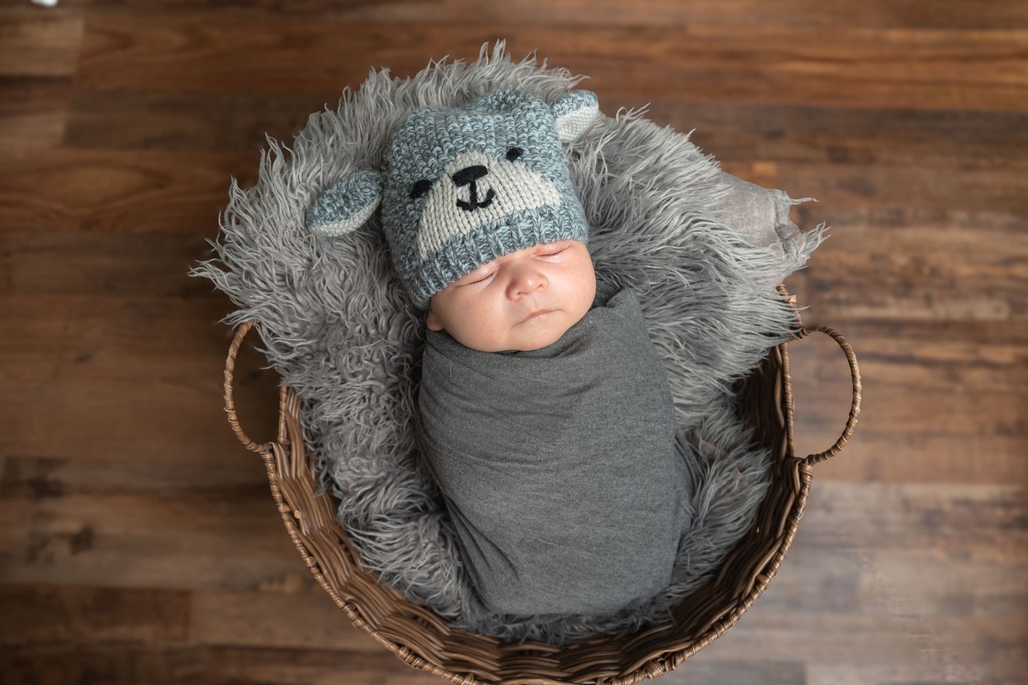 Newborn baby wrapped in gray swaddle wearing a knit bear hat, sleeping in a wicker basket with fluffy gray fur lining.