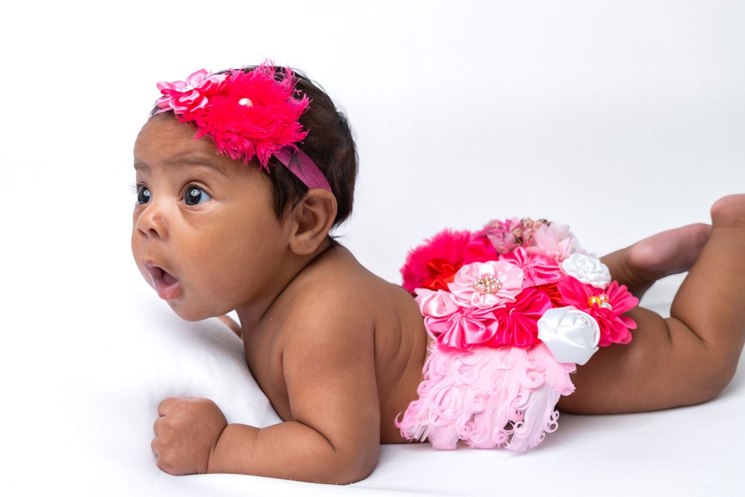 Adorable baby girl in pink floral headband and ruffled bloomers lying on white background.