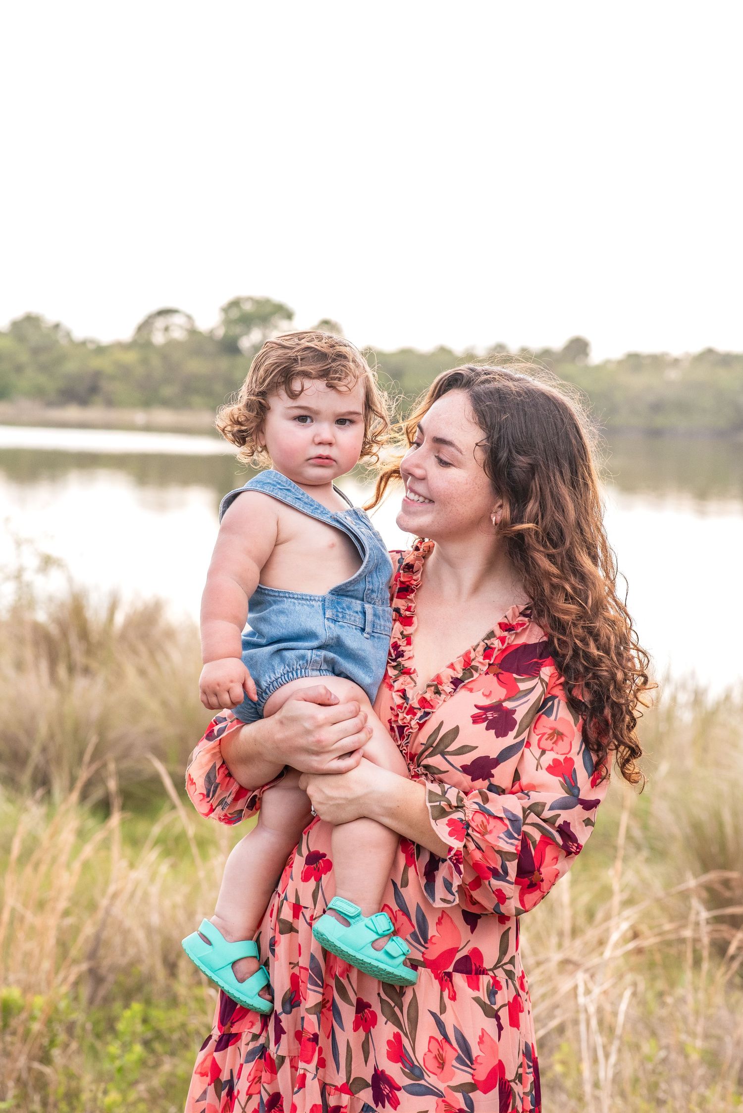 A smiling mother in a floral dress holds her toddler in denim overalls near a serene lake outdoors.