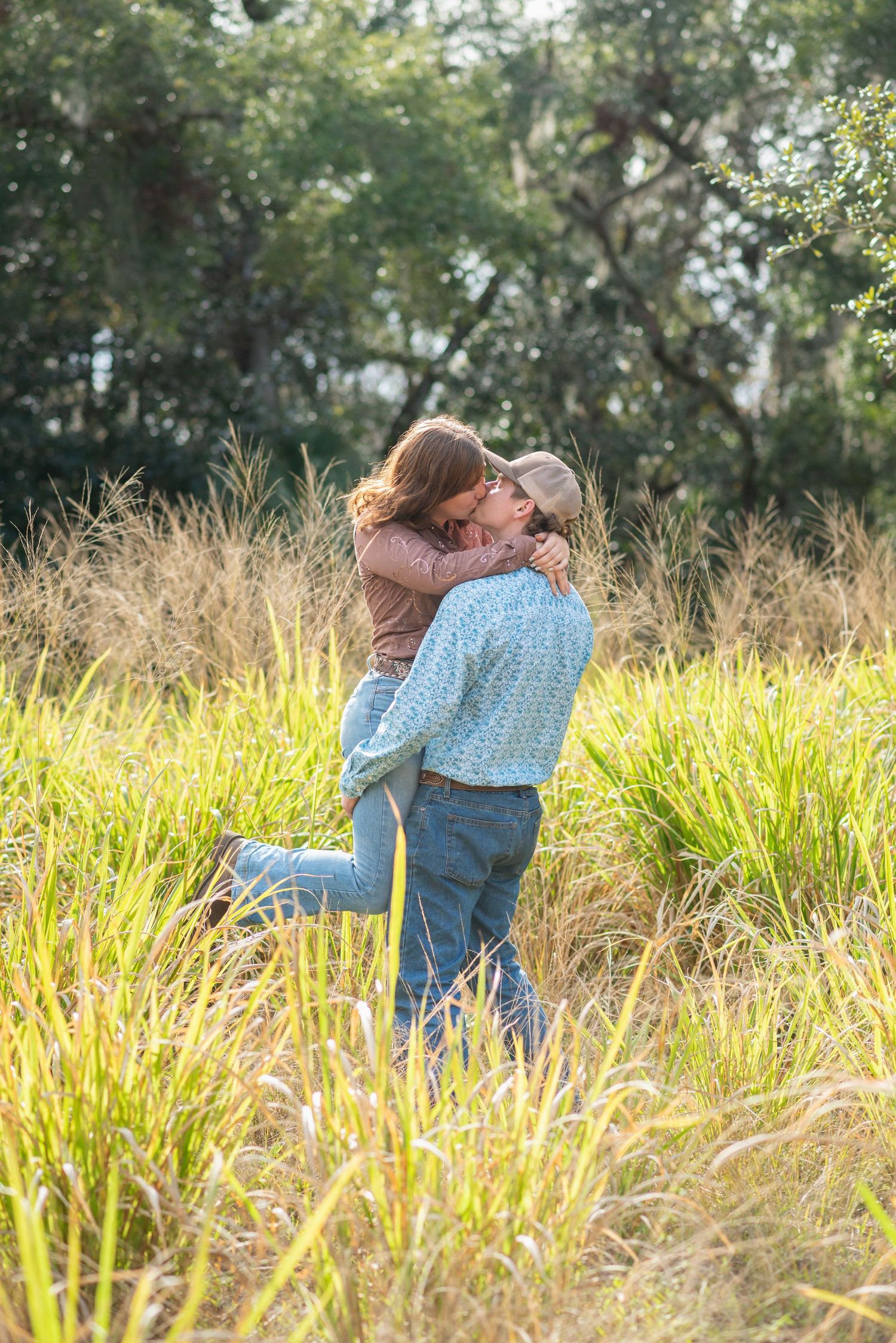 Couple embracing and kissing in a sunlit field of tall golden grass with lush green trees in the background.