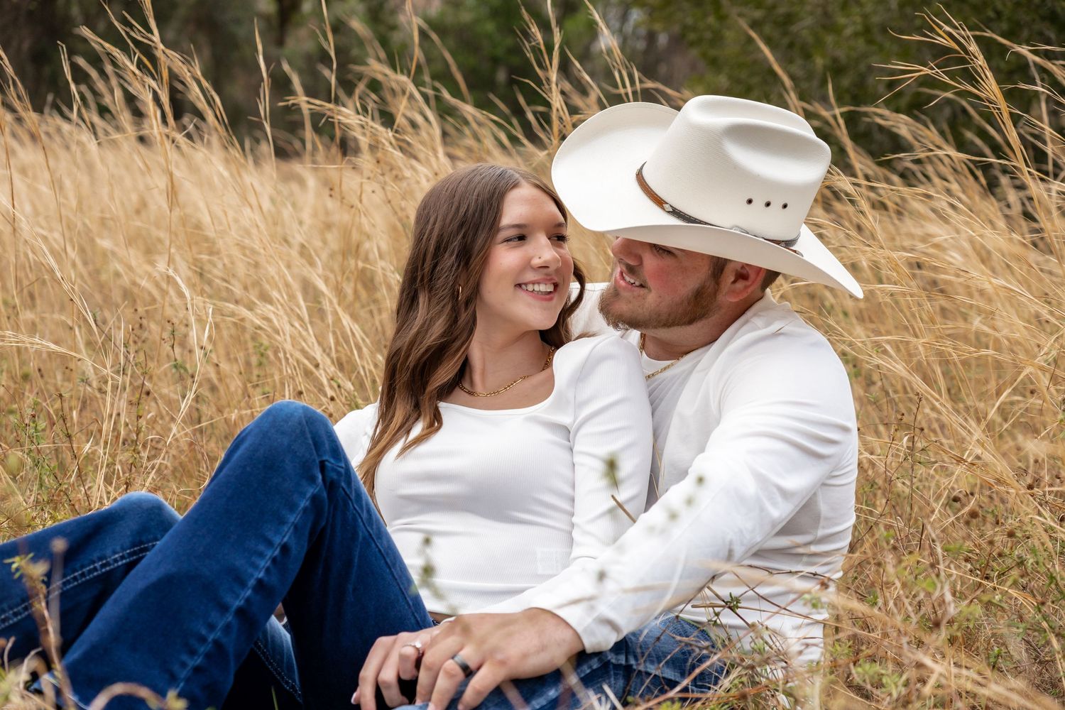 Couple sitting in golden grass field, woman in white top and jeans, man in white cowboy hat and shirt, smiling romantically.