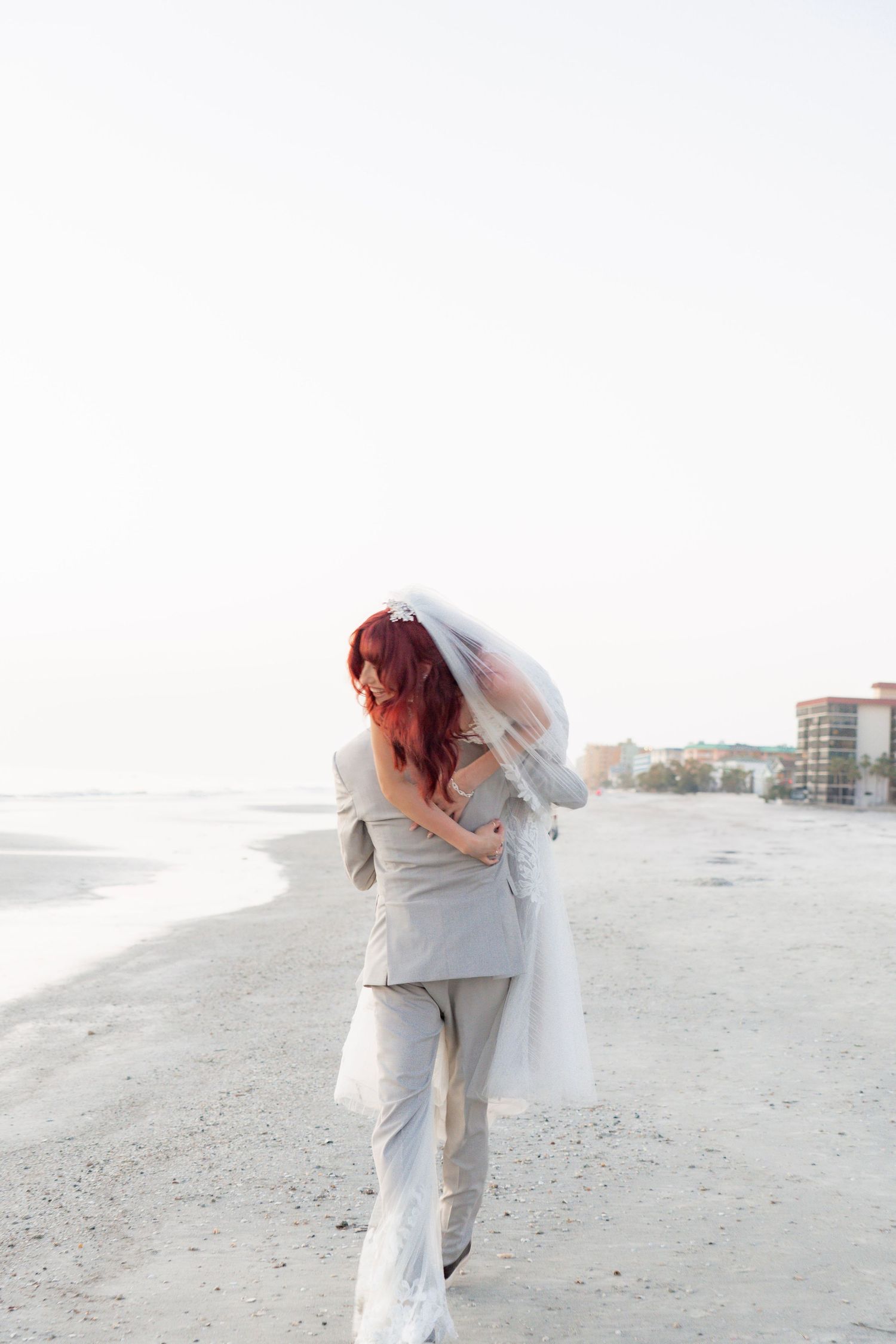 Bride with red hair and groom embracing on a beach, bride wearing a veil, in a romantic wedding photo.