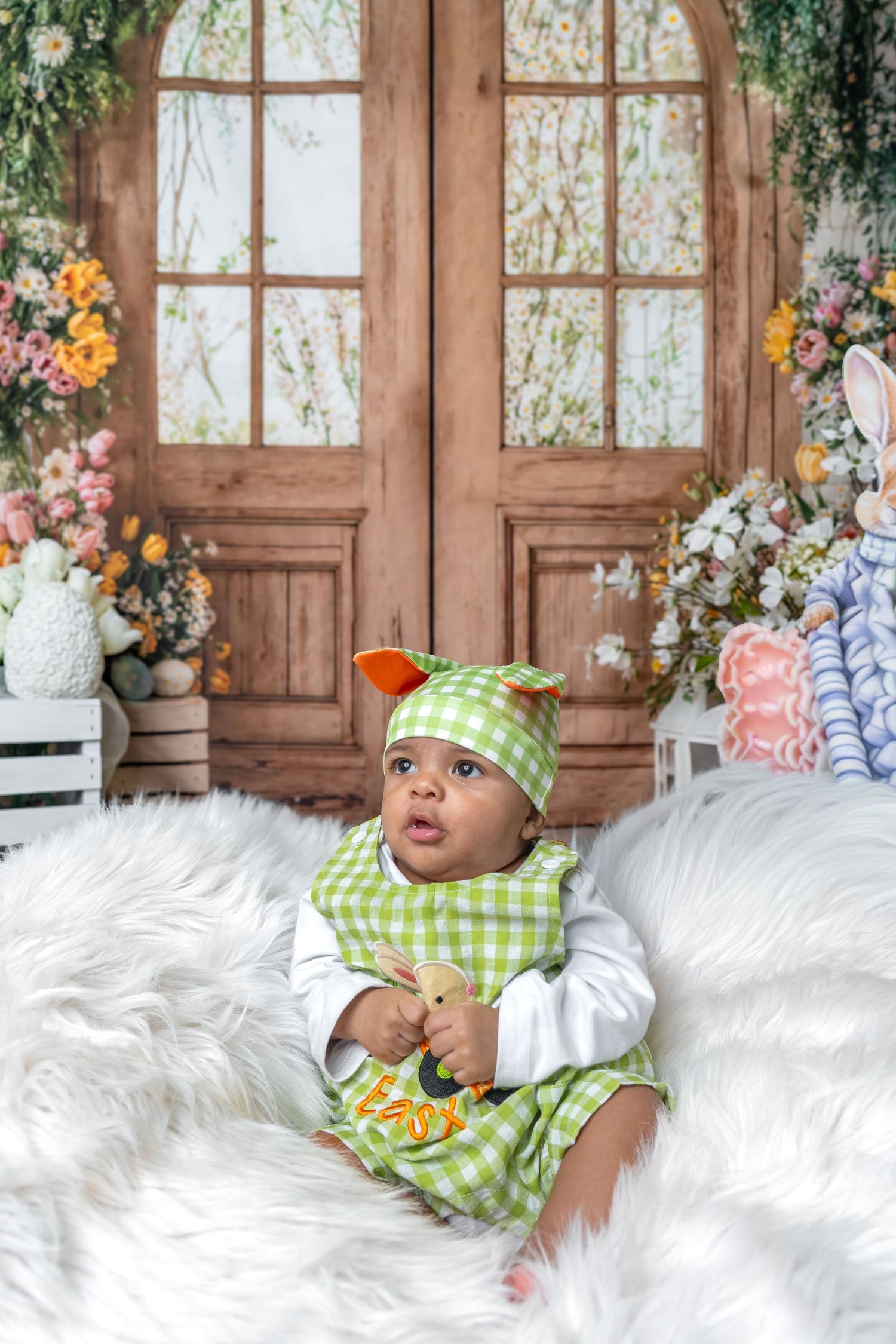 Baby in green gingham Easter outfit and hat sits on fluffy white fur, with floral door backdrop and bunny decor.