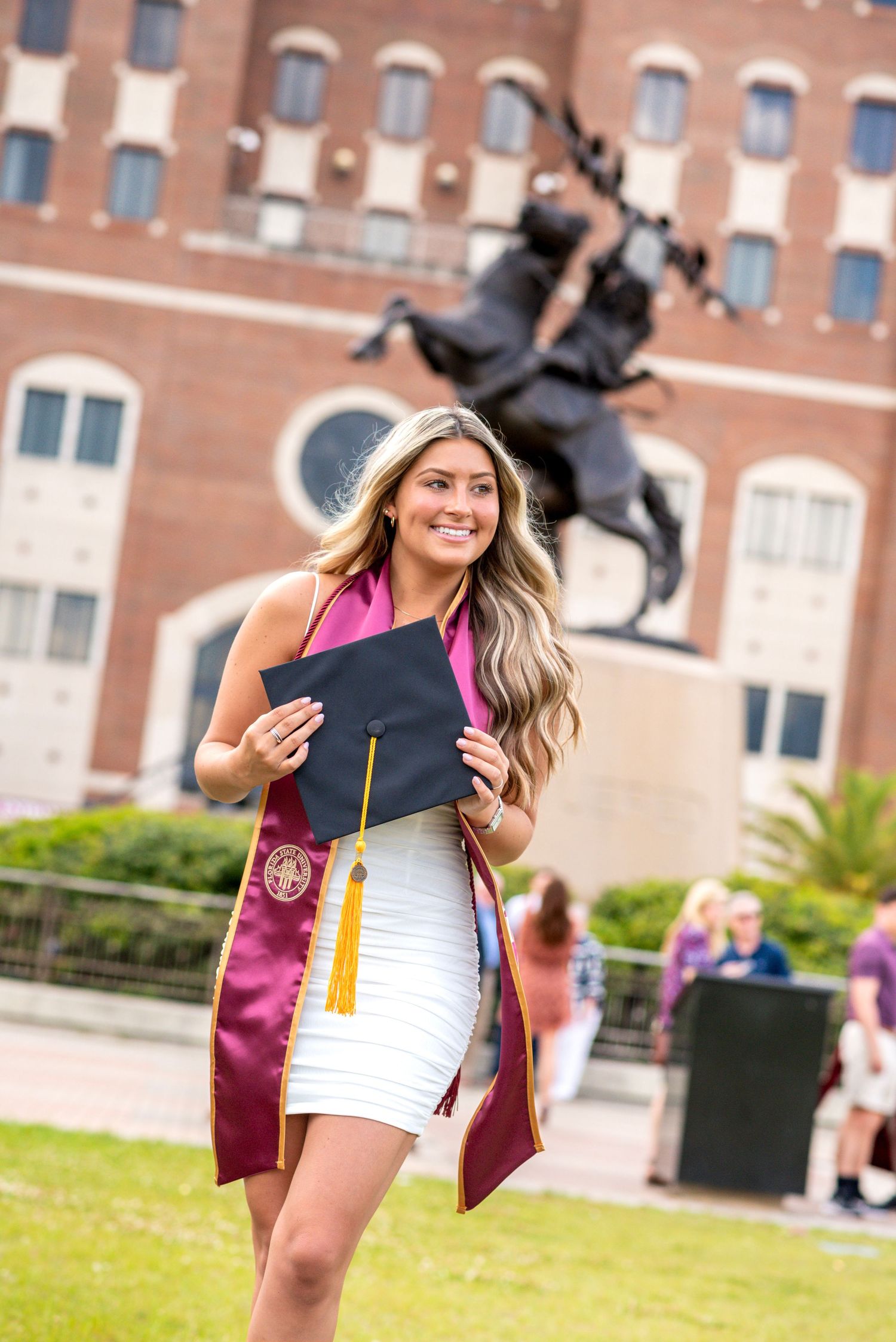 Young woman in white dress holding graduation cap with maroon stole, posing in front of university statue.