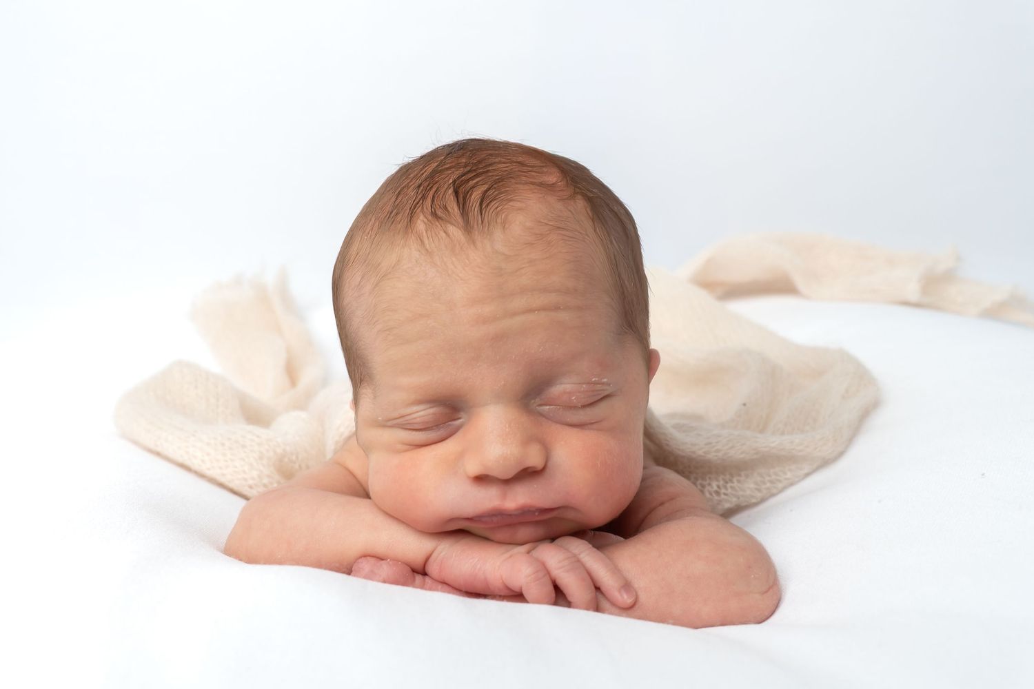 Sleeping newborn baby resting chin on hands, wrapped in a soft beige cloth on white background.