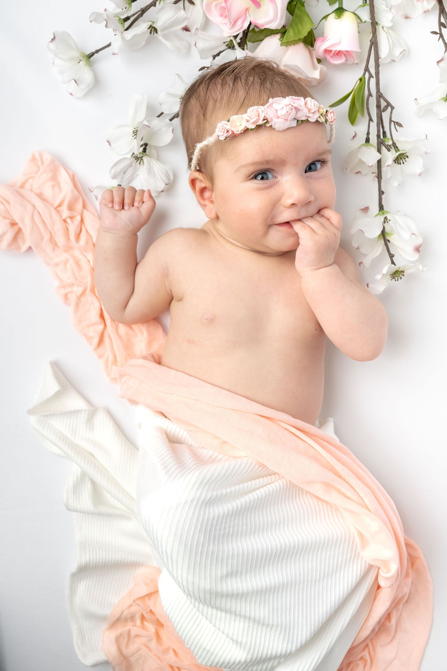 Adorable baby girl lying among pink and white flowers, wearing a floral headband, with a peach fabric wrap.