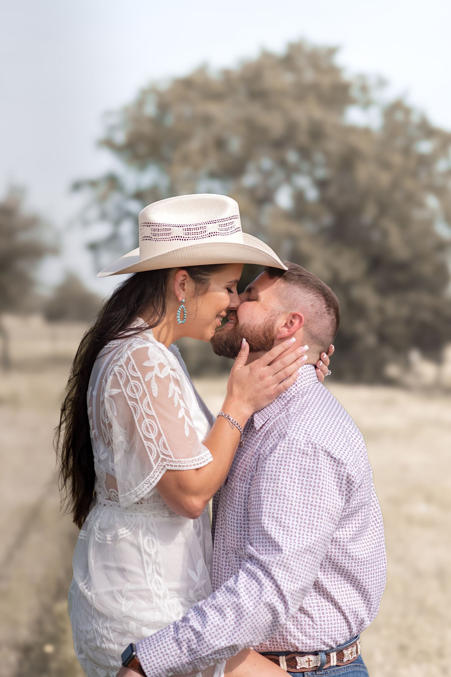 A couple shares a romantic kiss outdoors, woman wearing a white cowboy hat and lace dress, man in a plaid shirt.