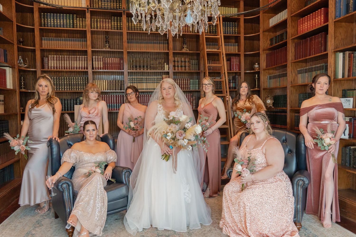 Bride in white gown poses with bridesmaids in pink and mauve dresses in an elegant library with bookshelves and chandelier.