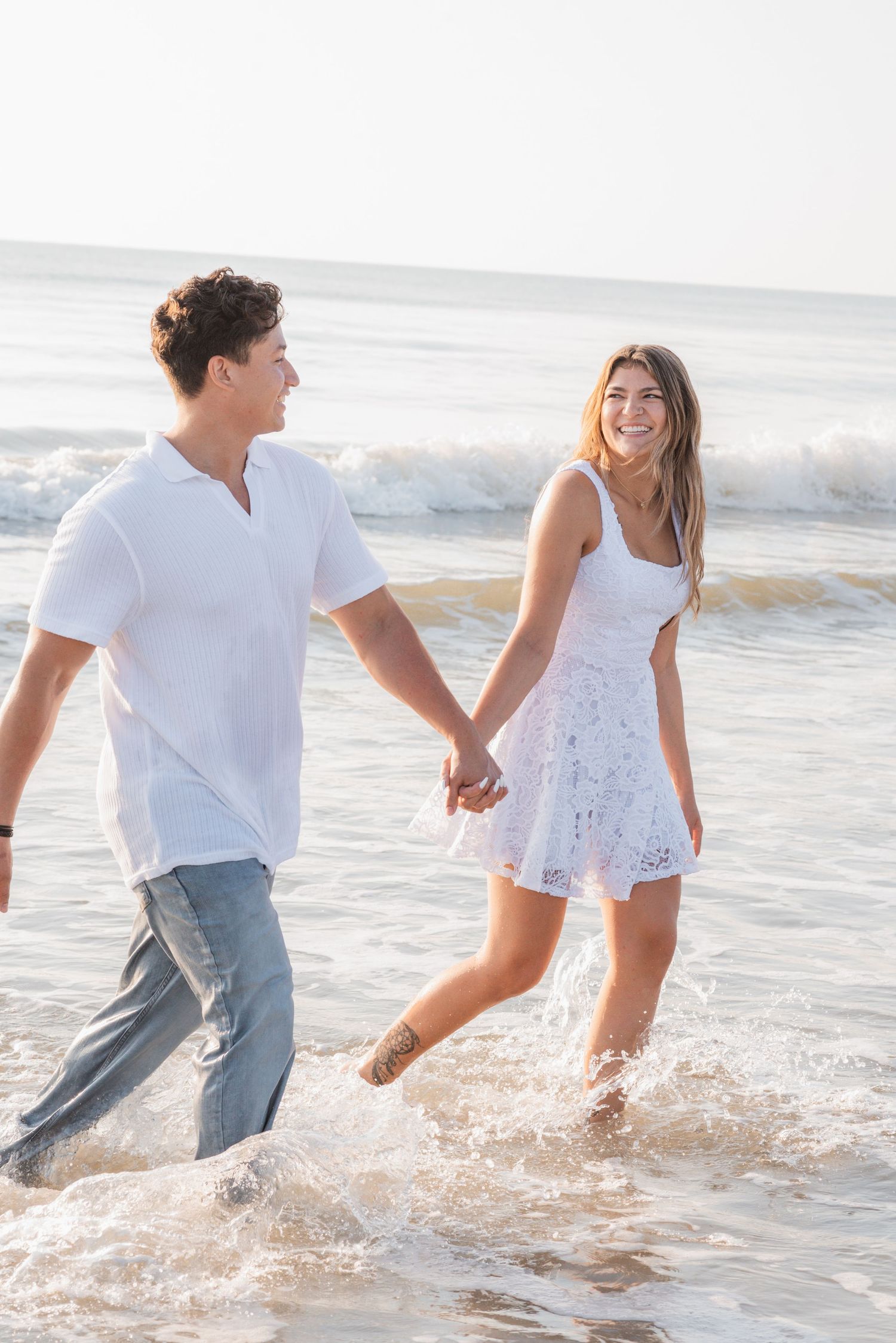 Happy young couple holding hands walking through ocean waves together.