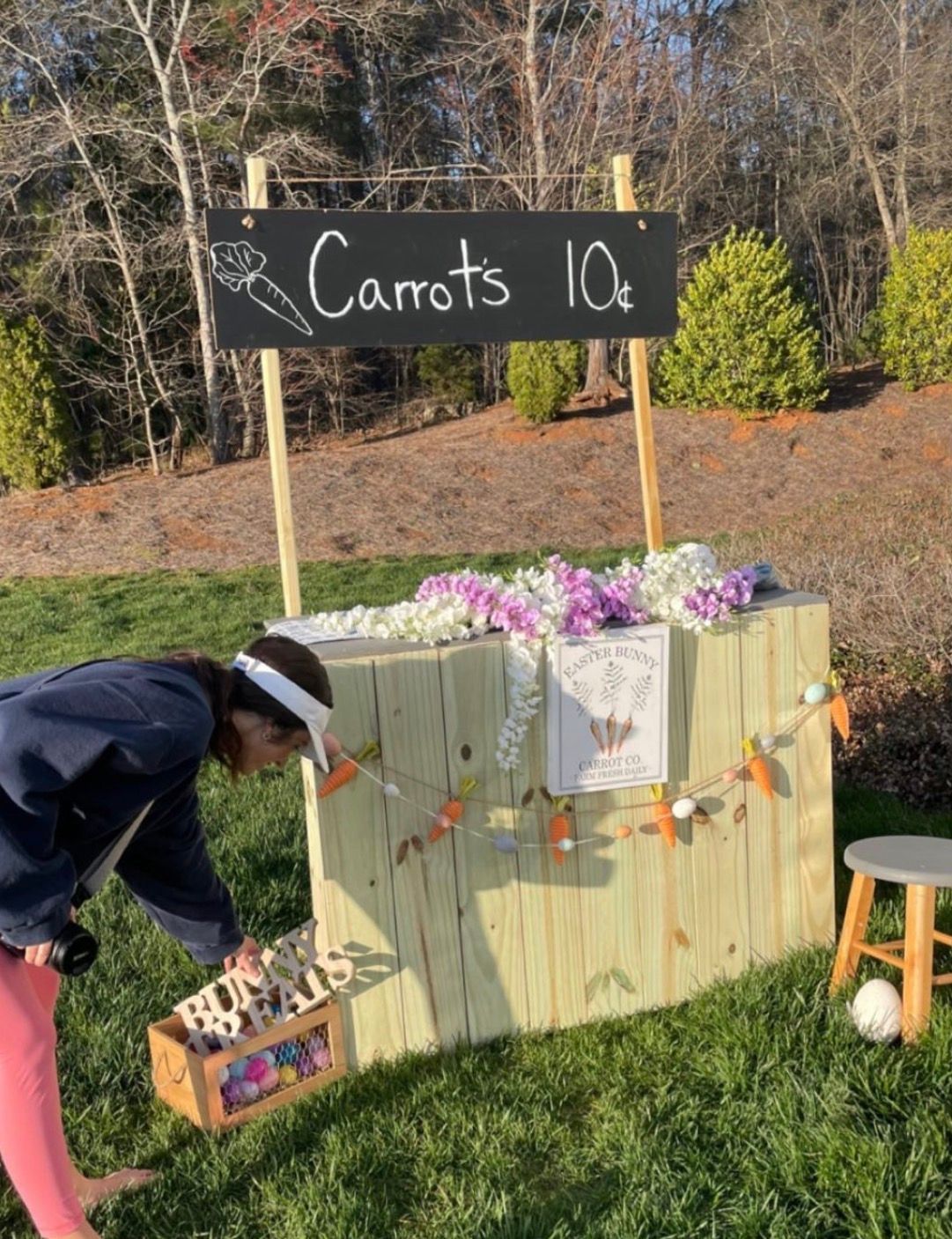 Child at a rustic wooden carrot stand selling carrots for 10 cents, decorated with flowers and bunting.