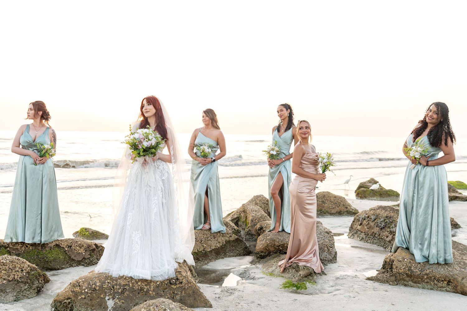 Bride in white gown with bridesmaids in mint and blush dresses holding bouquets on a rocky beach at sunset.