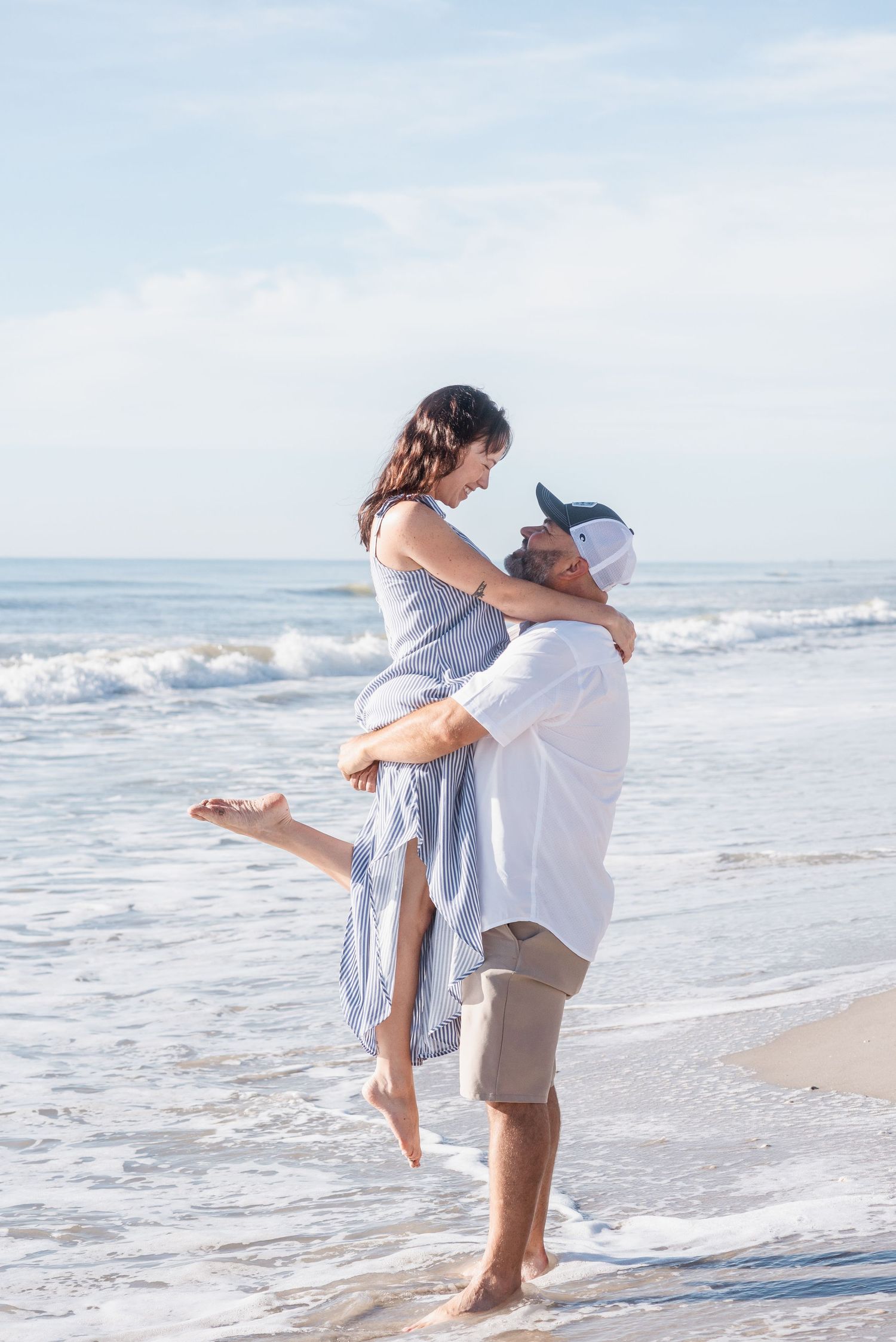 A man lifts a woman in his arms on a sunny beach, both smiling and gazing at each other near ocean waves.