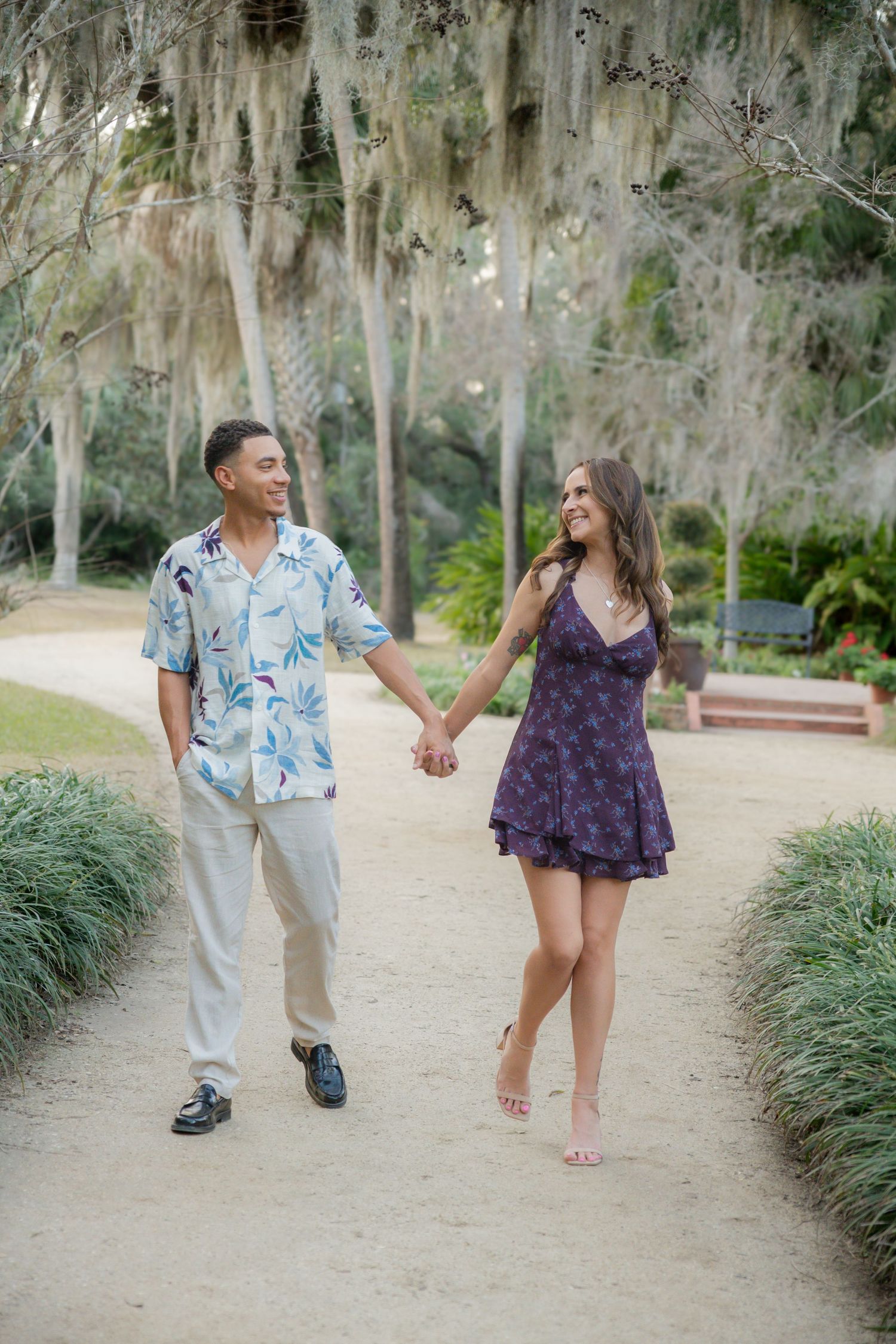Happy couple holding hands and walking along a scenic garden path lined with Spanish moss-draped trees.