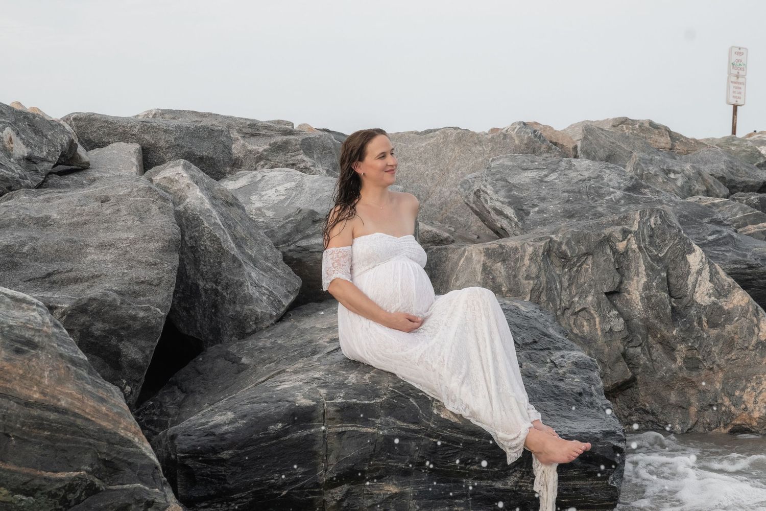 Pregnant woman in white dress sitting on rocky jetty by ocean for maternity photo shoot.