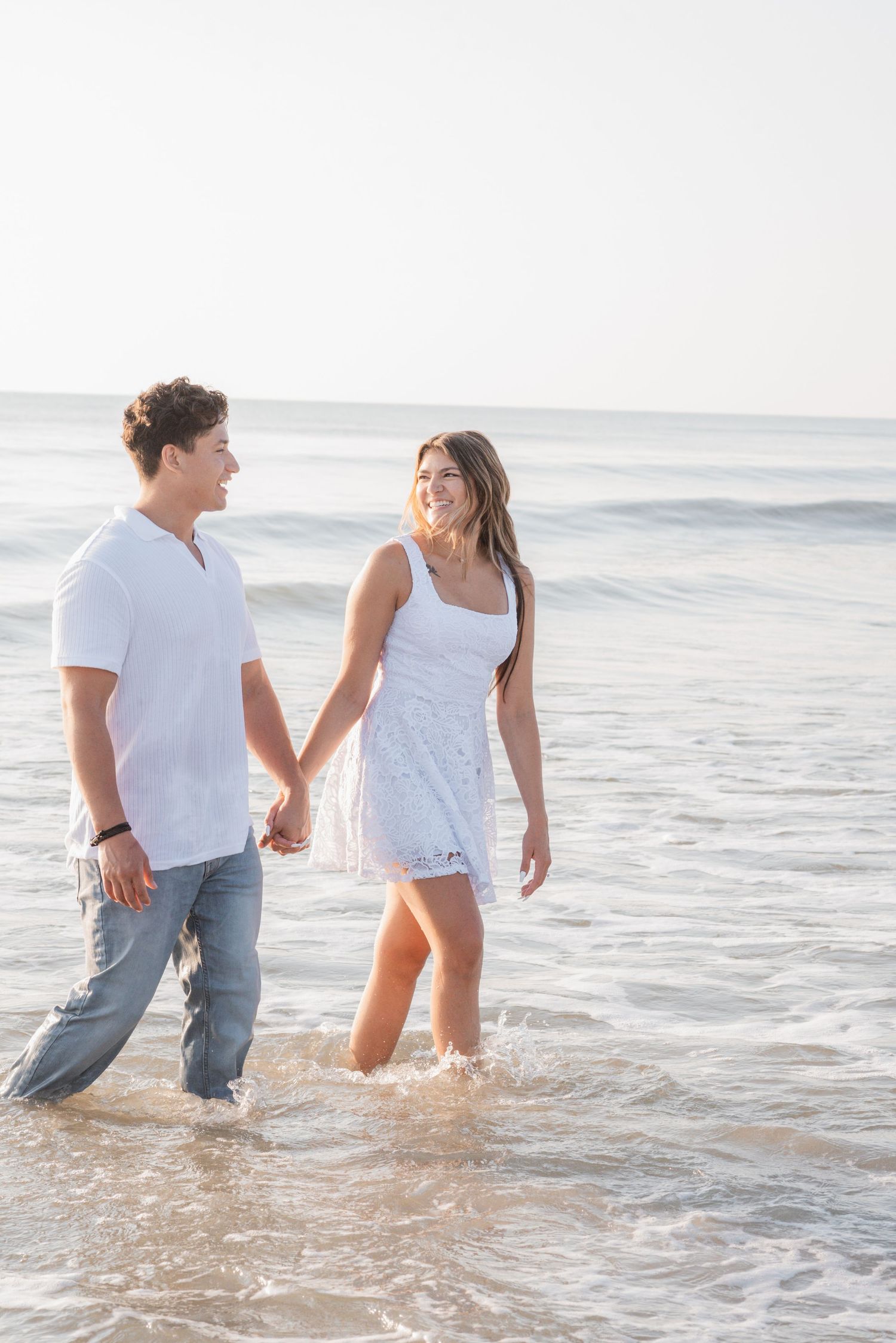 Romantic couple holding hands and walking through shallow ocean waves on a sunny beach in matching white outfits.