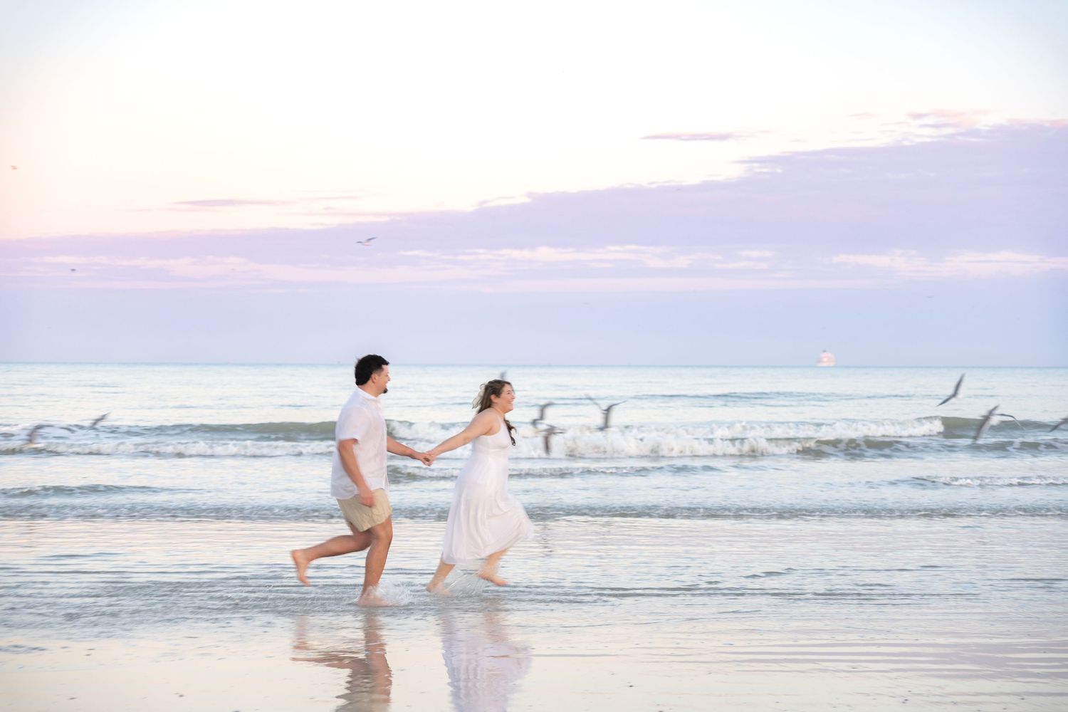 Couple running playfully along the shoreline at sunset, woman in white dress, seagulls flying in background.