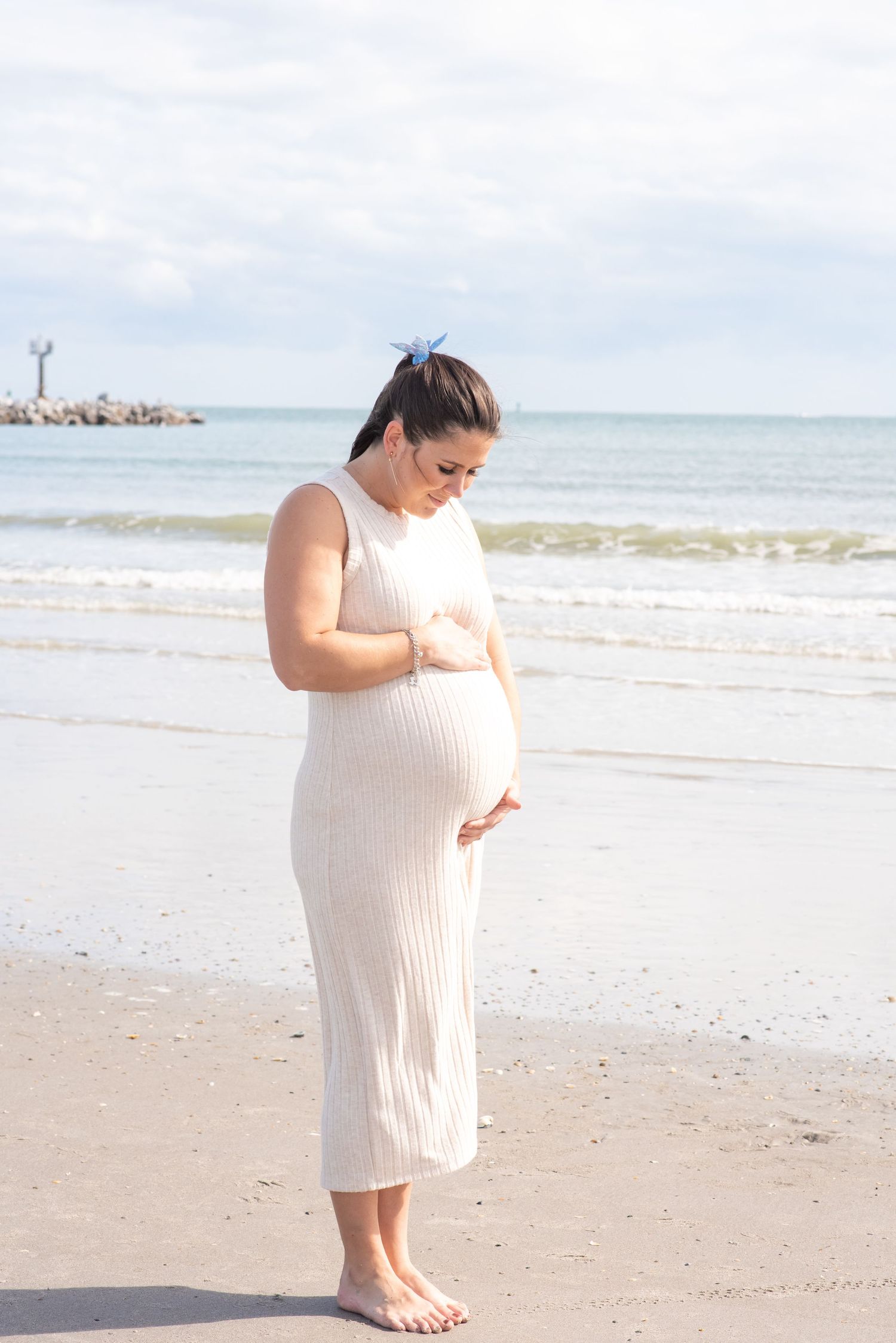 Pregnant woman in white ribbed dress cradling baby bump on sandy beach with ocean waves behind her.