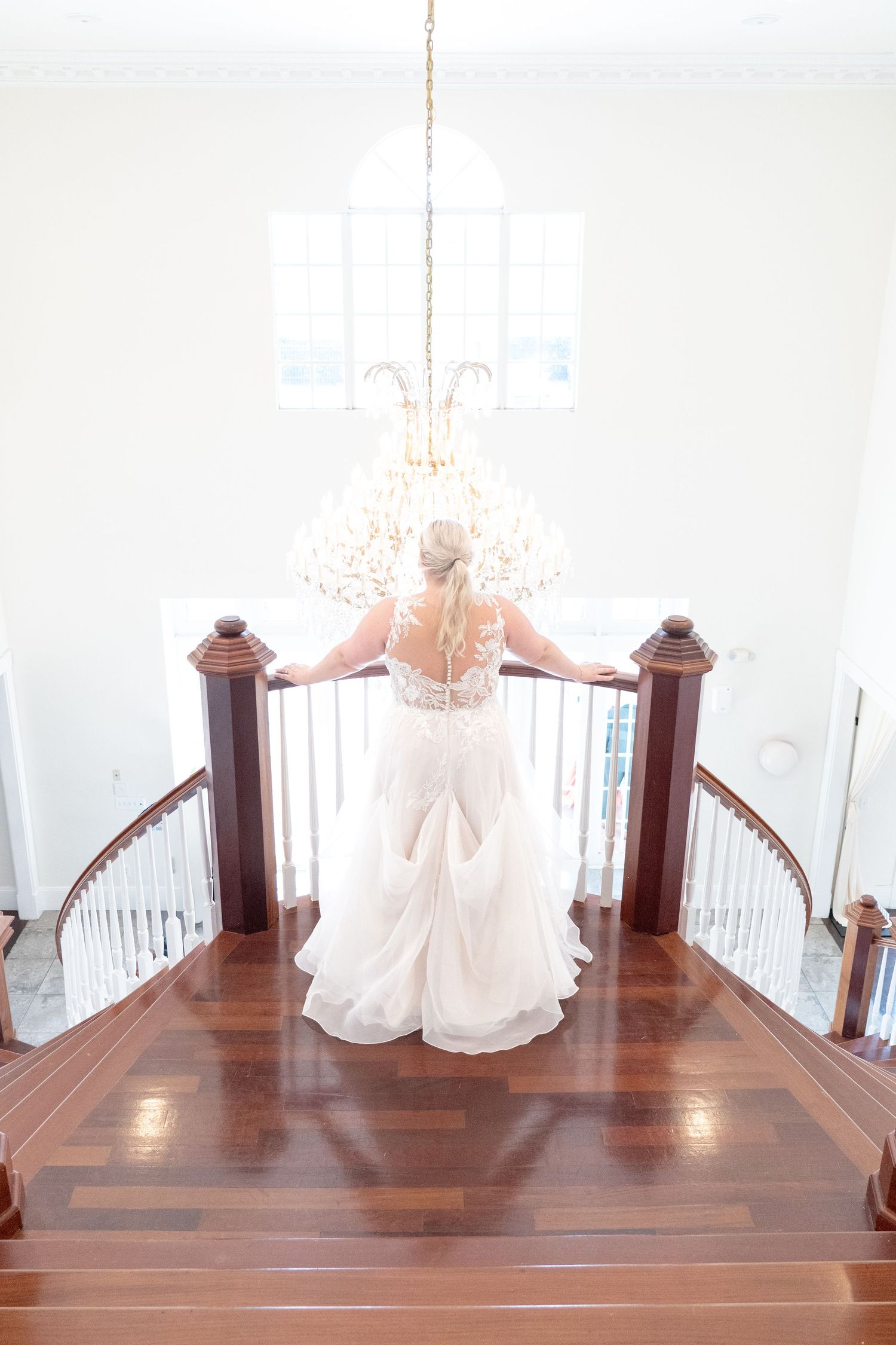 Bride in white lace gown stands atop elegant curved staircase beneath crystal chandelier in bright wedding venue.