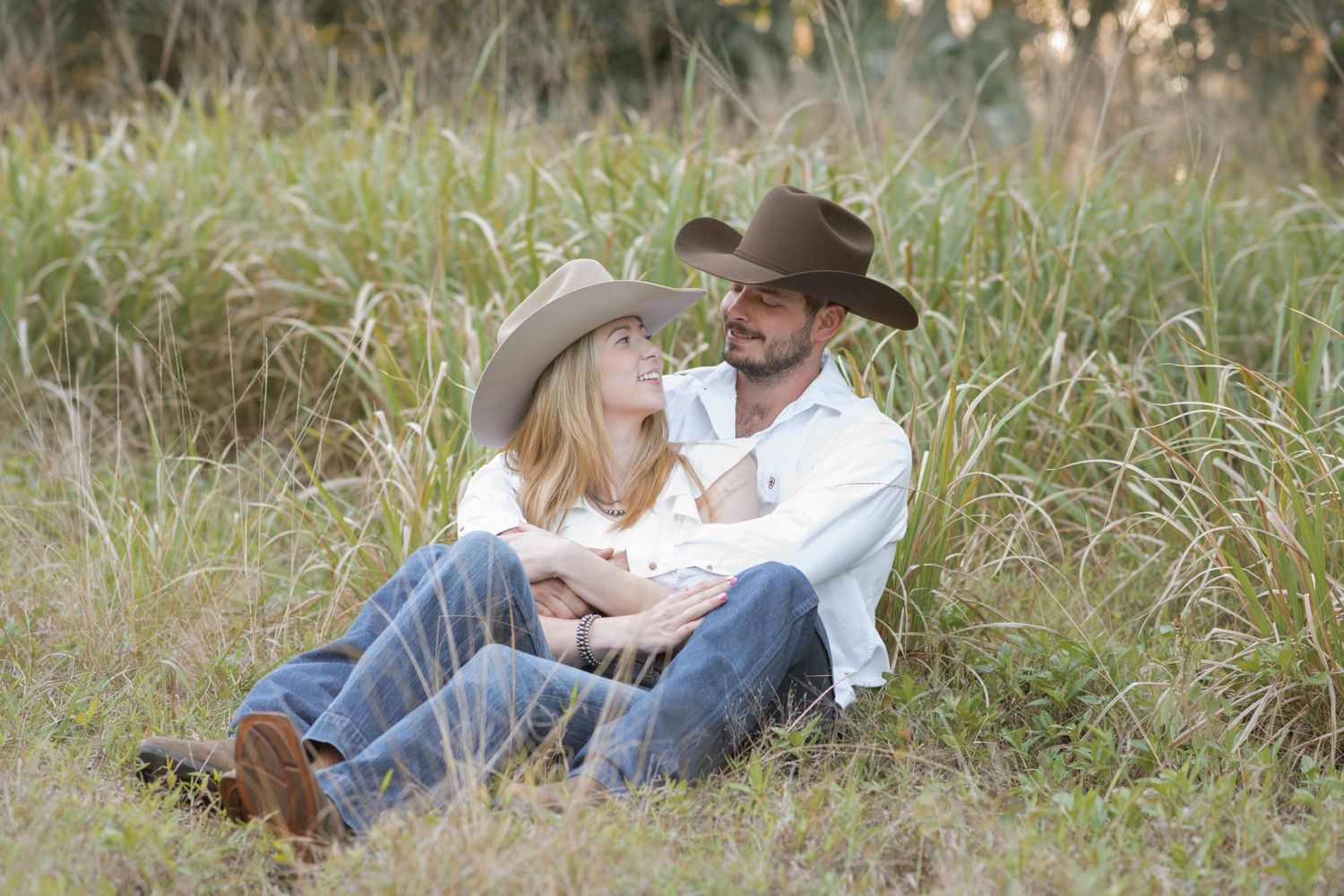 Couple in cowboy hats sitting together in tall grass field, sharing a romantic moment outdoors.