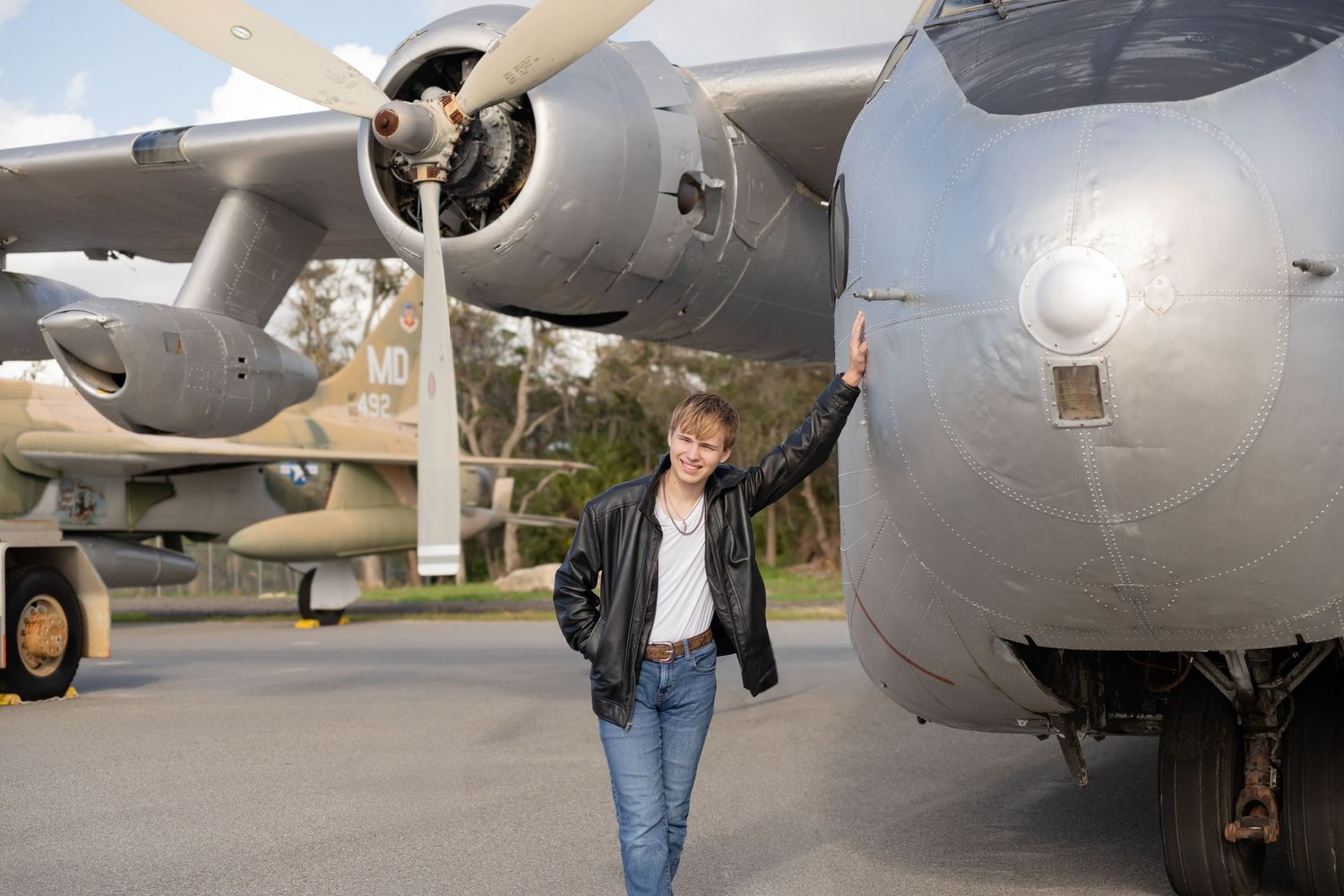 Woman in leather jacket standing beside a large military aircraft propeller engine outdoors.