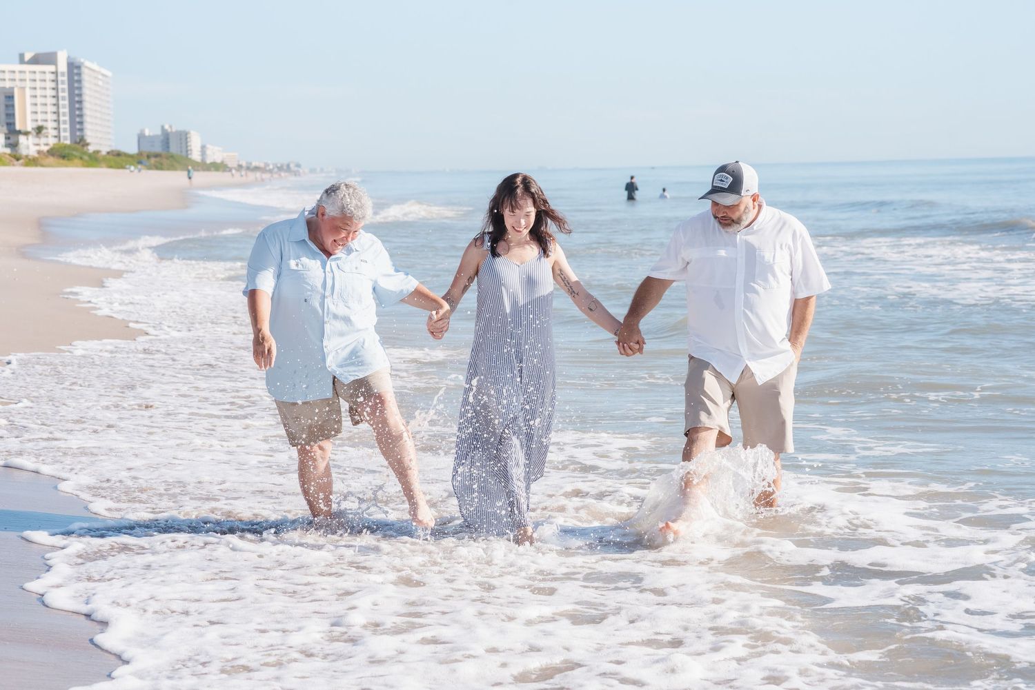 Three people laugh and splash in ocean waves on a sunny beach, holding hands and enjoying the water together.
