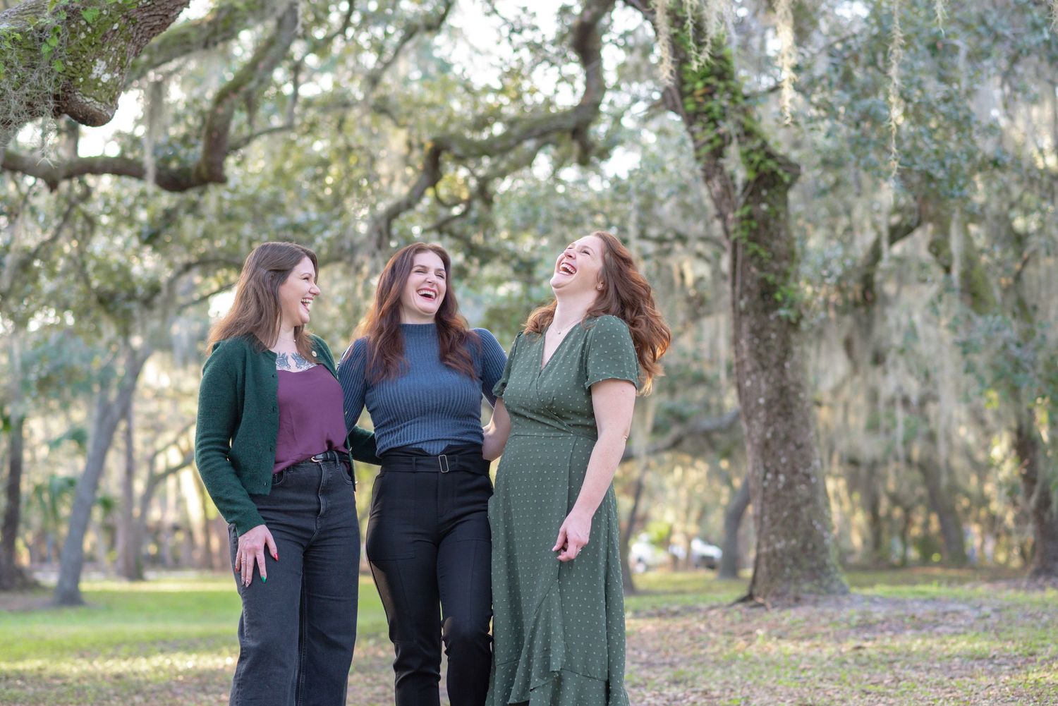 Three women laughing and walking together outdoors under moss-covered oak trees in a lush green park setting.