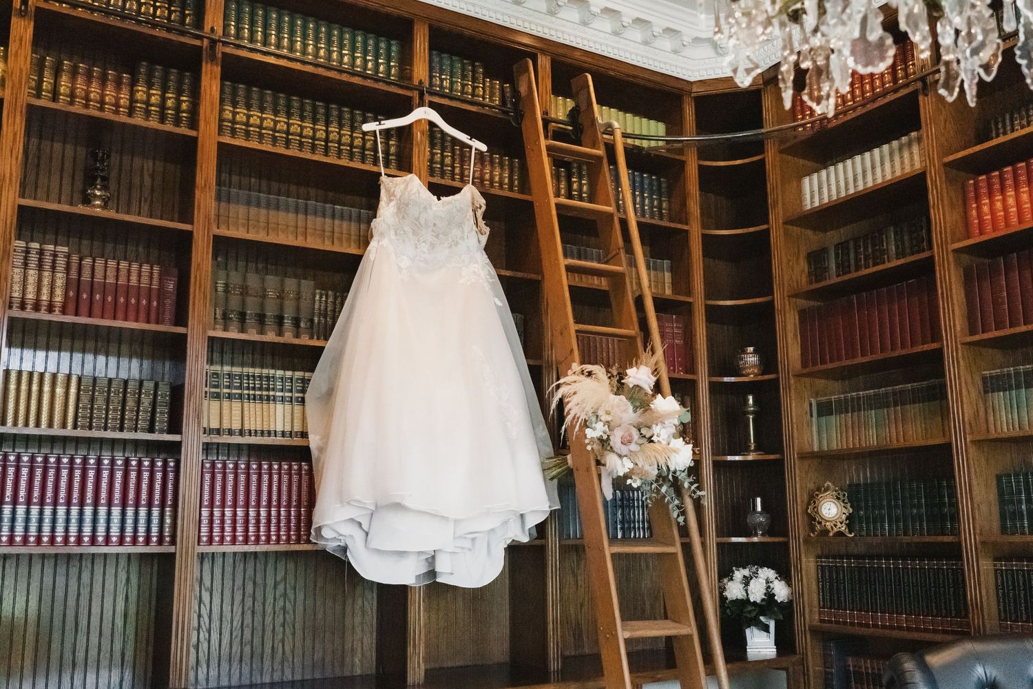 White wedding dress hanging on a ladder in an elegant library with floor-to-ceiling bookshelves and a bridal bouquet.