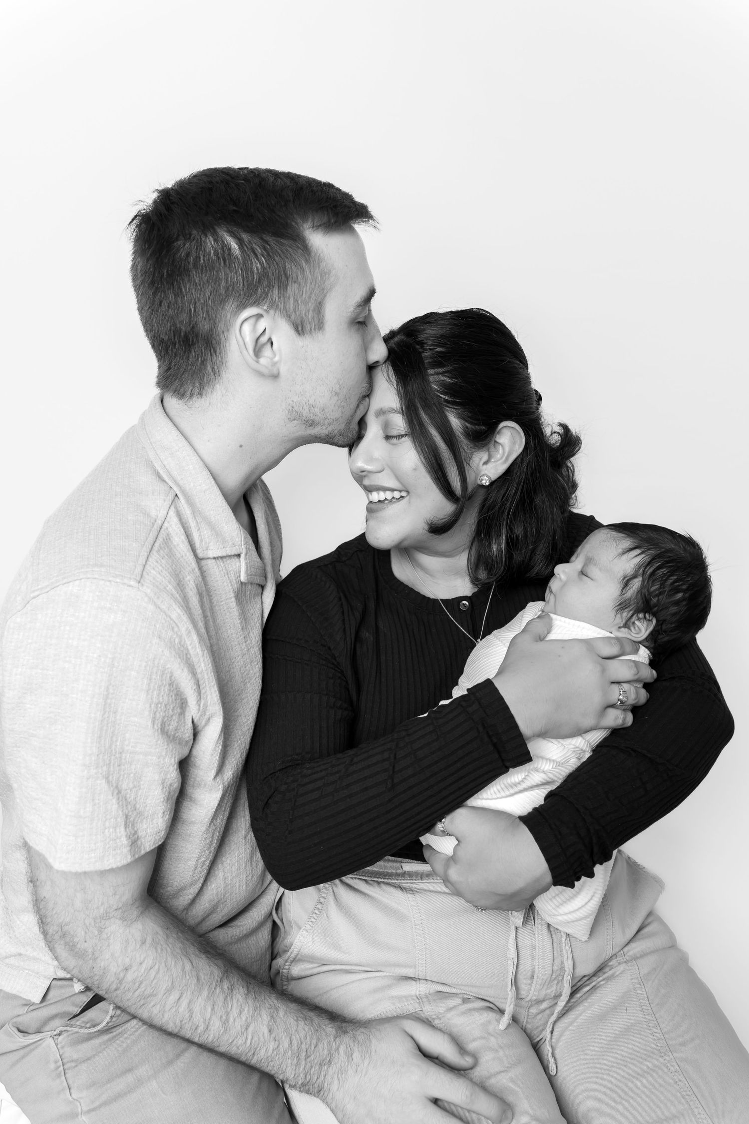 Black and white photo of a couple smiling and kissing while tenderly holding their newborn baby.