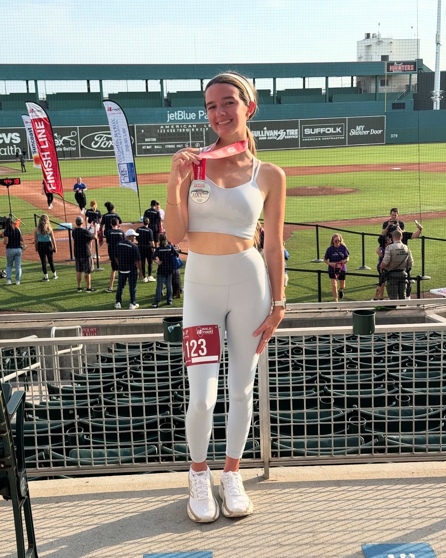 Athletic woman in white workout set holding a race medal at JetBlue Park baseball stadium after a run.