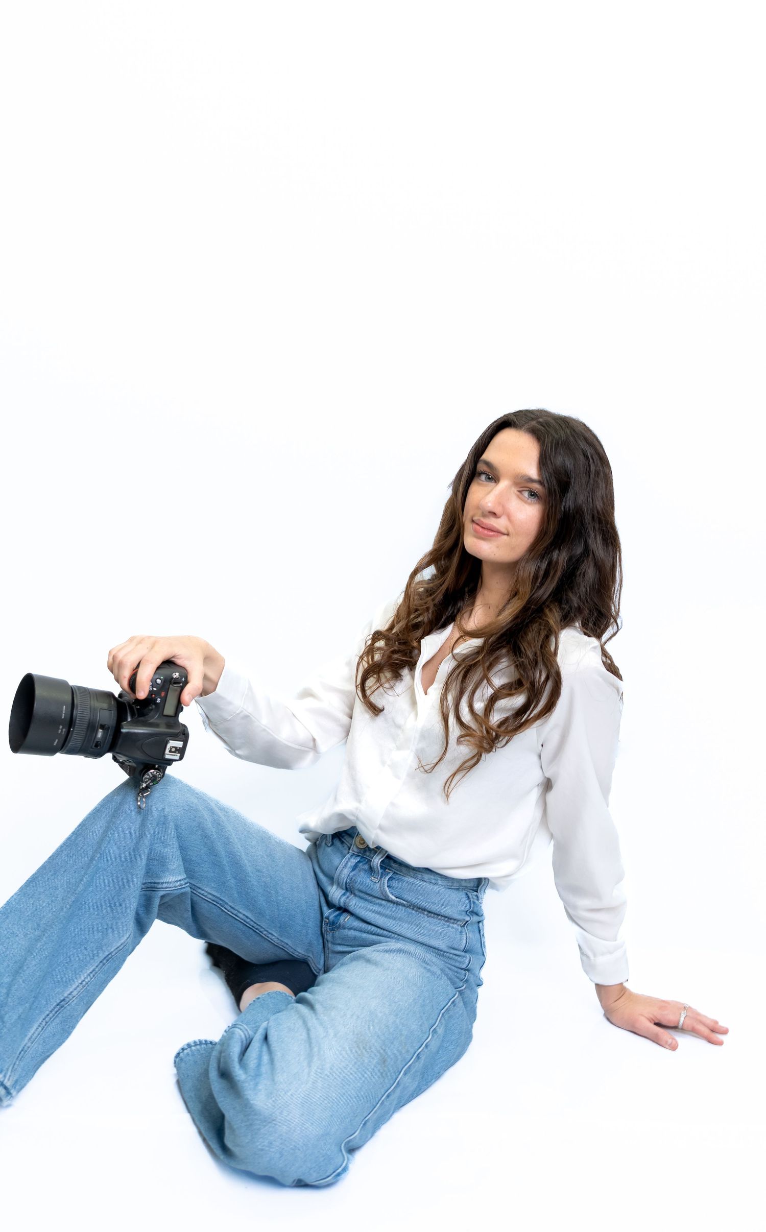 Female photographer sitting on white floor holding DSLR camera, wearing white blouse and blue jeans, smiling.