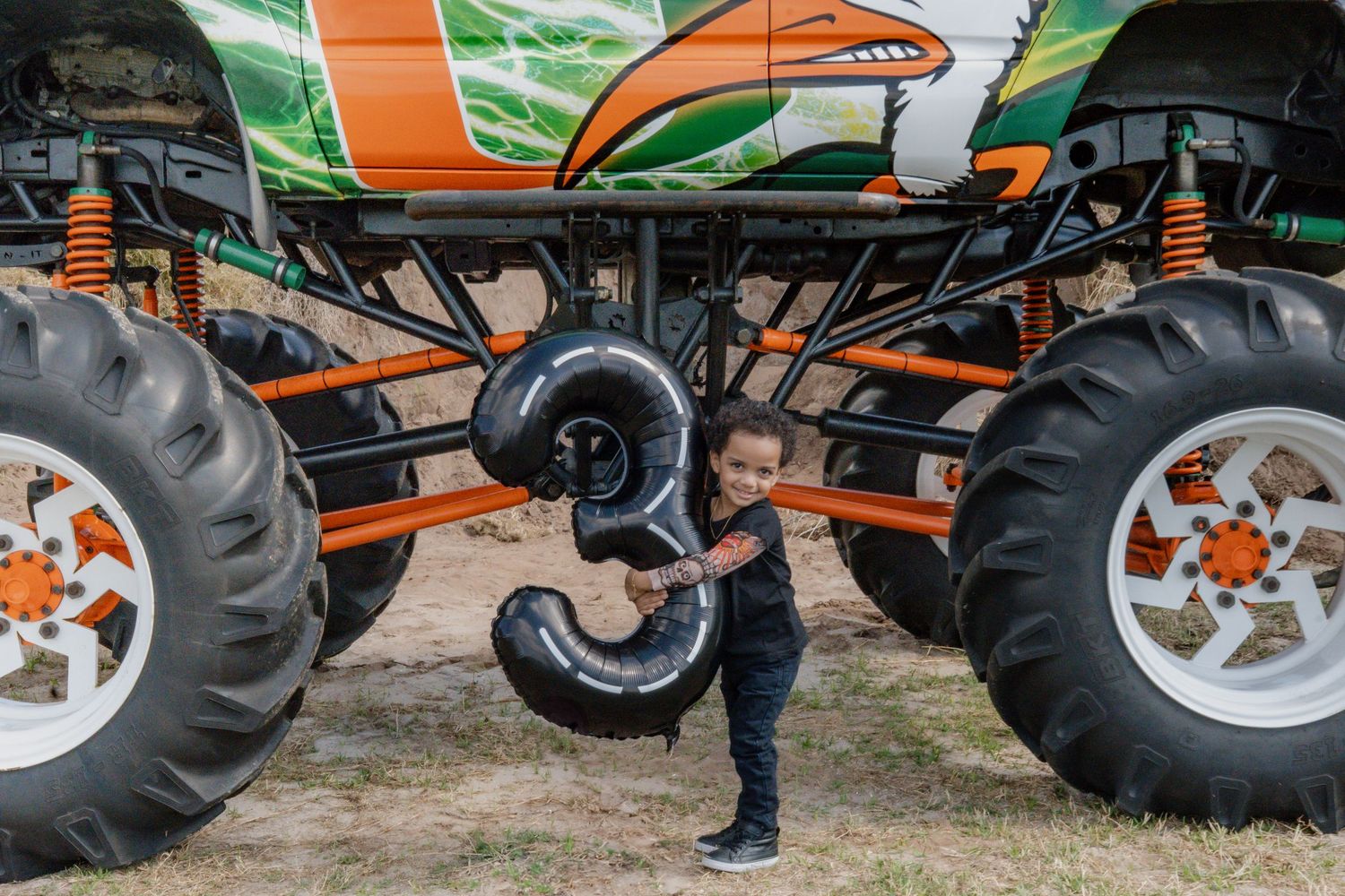 Toddler boy holding number 3 balloon standing under colorful monster truck chassis.
