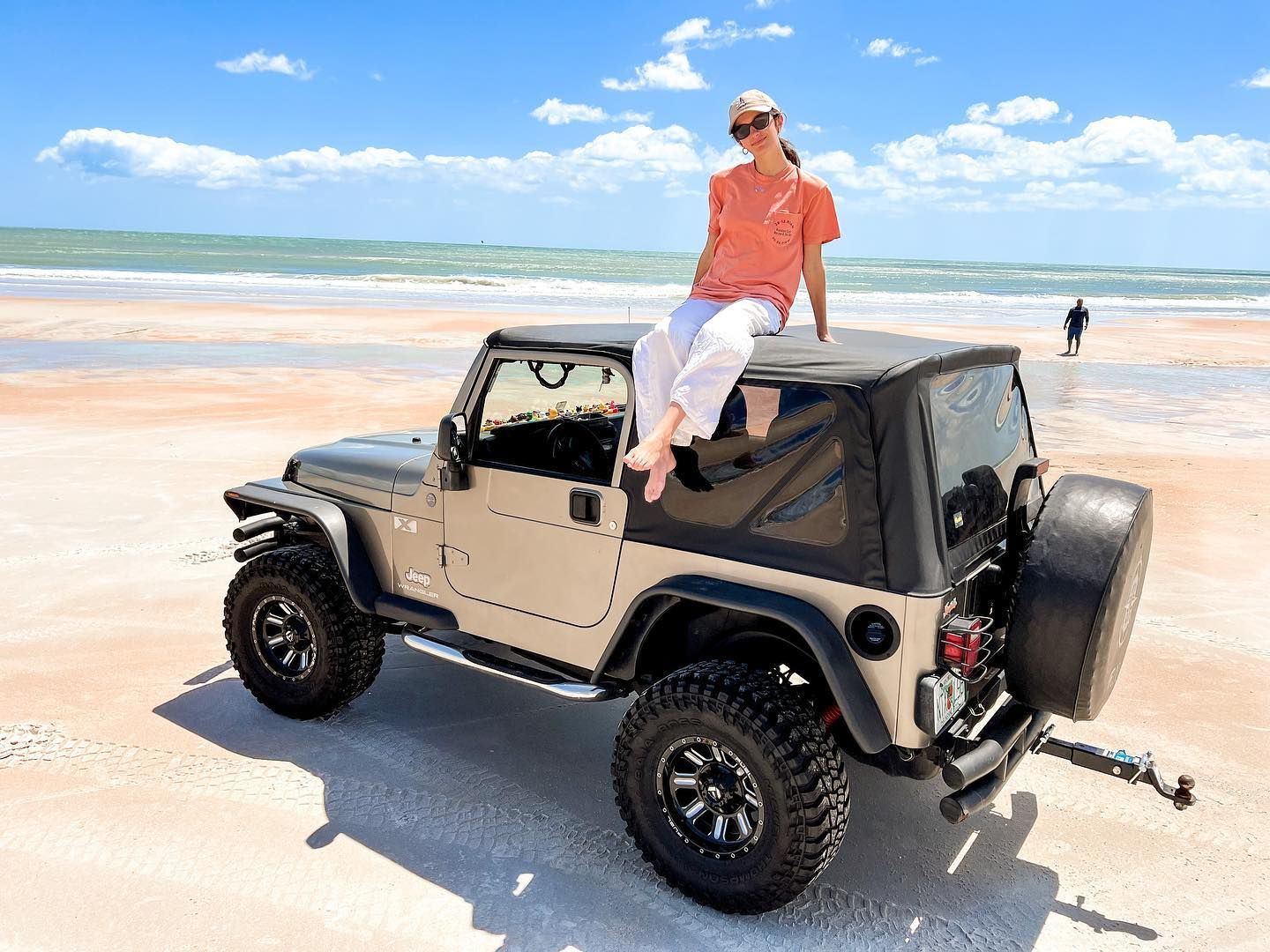 Woman sitting on top of a tan Jeep Wrangler on a sandy beach with blue ocean waves in the background.