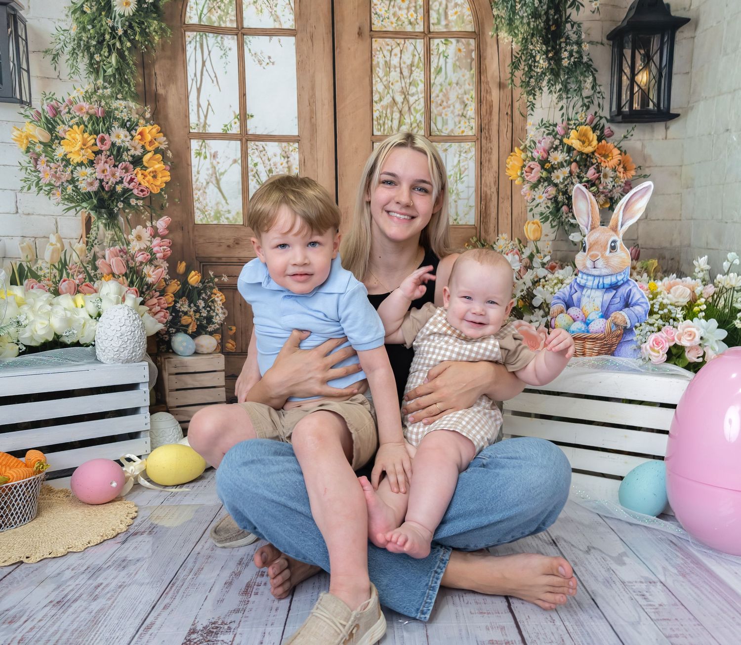 Young woman sits cross-legged holding two toddlers in an Easter-themed photo with colorful eggs, flowers, and bunny decor.