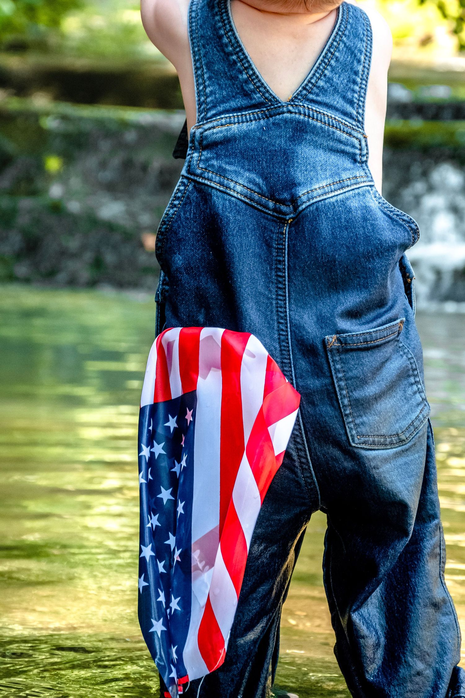 Child in denim overalls with American flag tucked in pocket, standing near water outdoors.