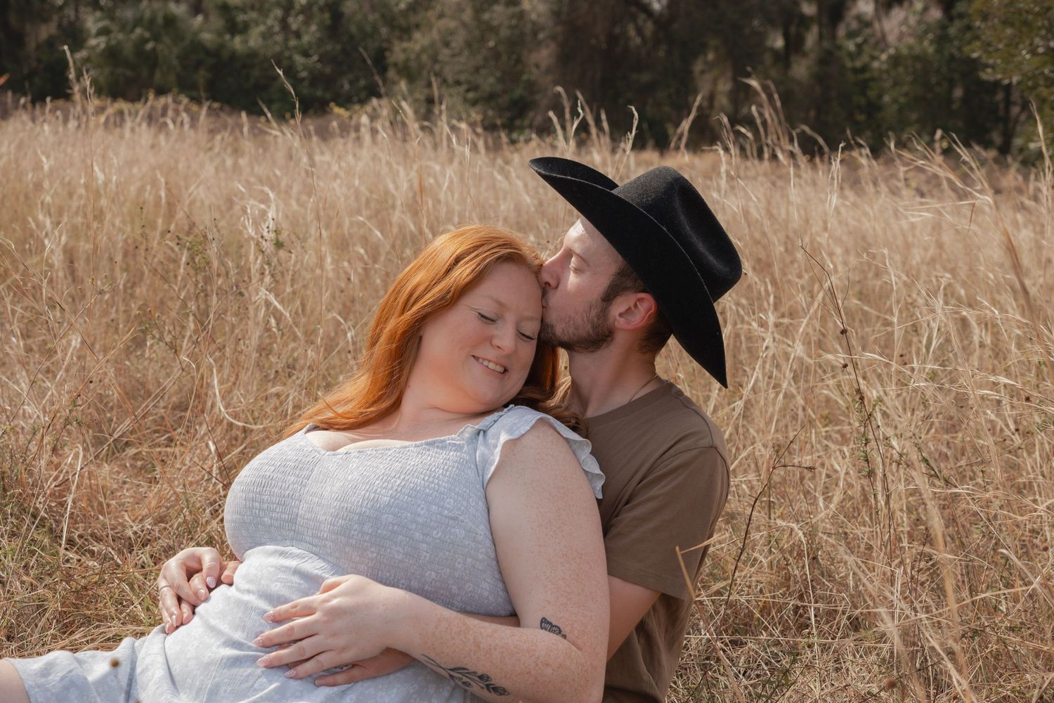 A man in a cowboy hat kisses a redheaded woman while sitting together in a golden field.