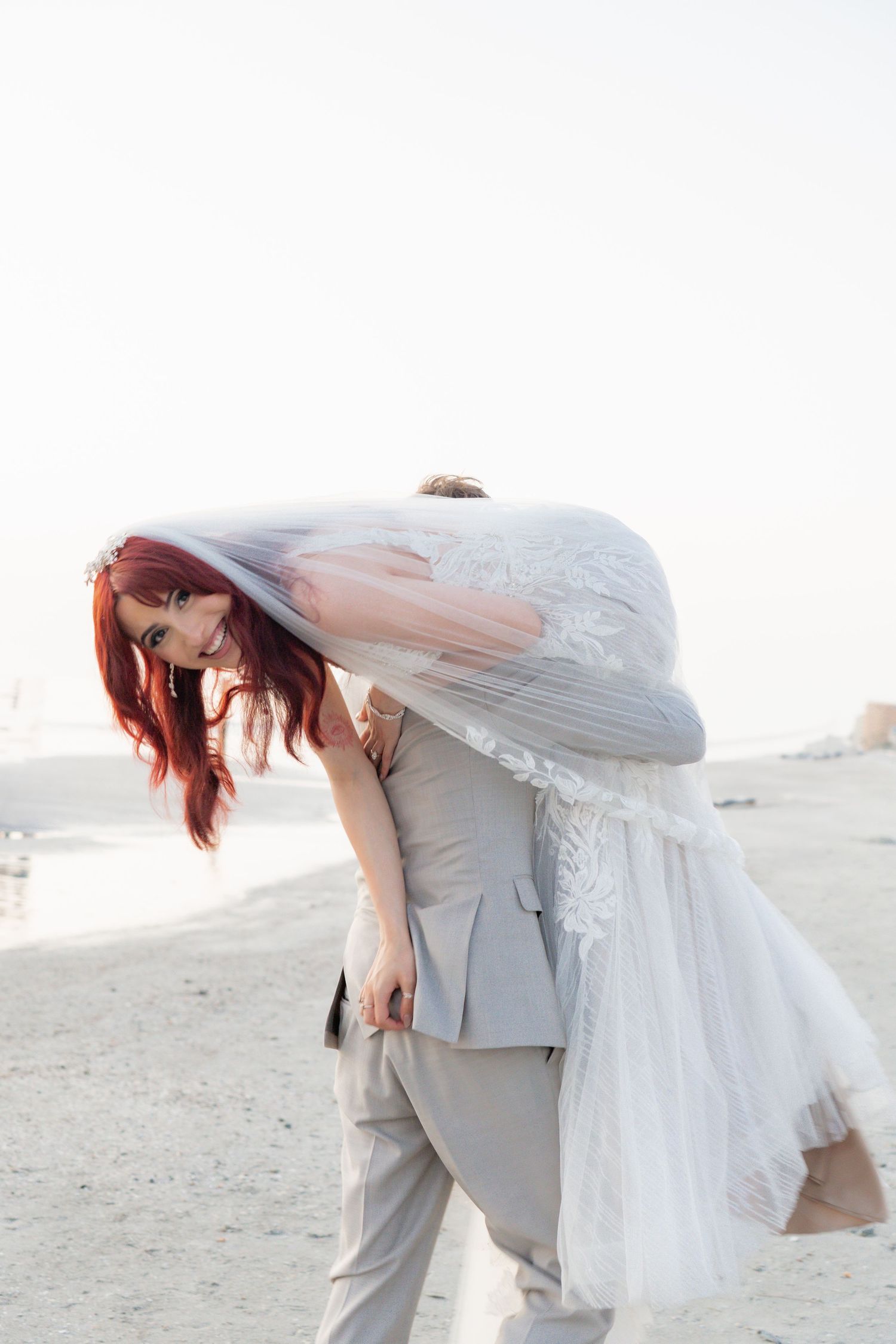 Groom carrying bride in white lace wedding dress over his shoulder on sandy beach, bride smiling with red hair.