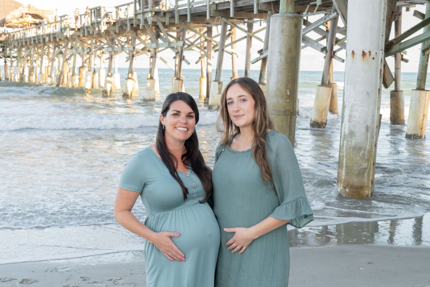 Two pregnant women in matching sage green dresses posing together under a beach pier.