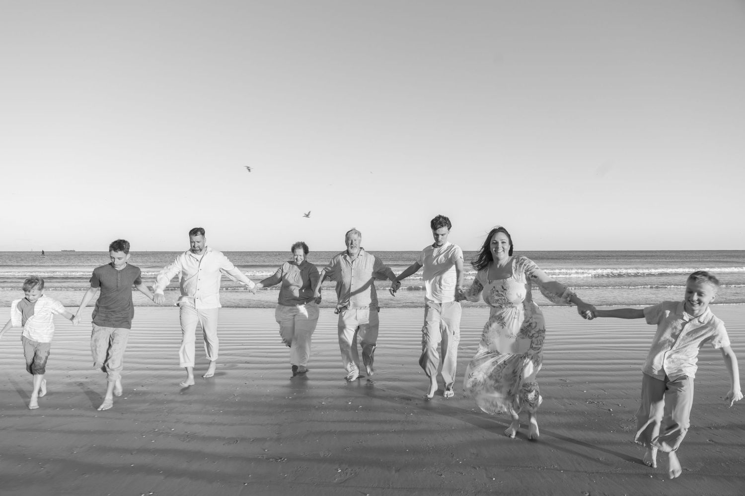 Black and white photo of a large family holding hands and running together on a beach near the ocean.