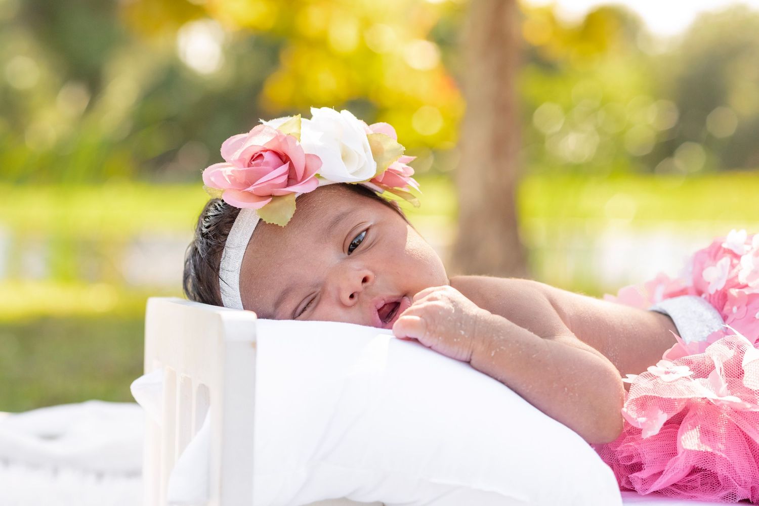 Newborn baby girl wearing a pink floral headband lying on a white pillow in an outdoor garden setting.