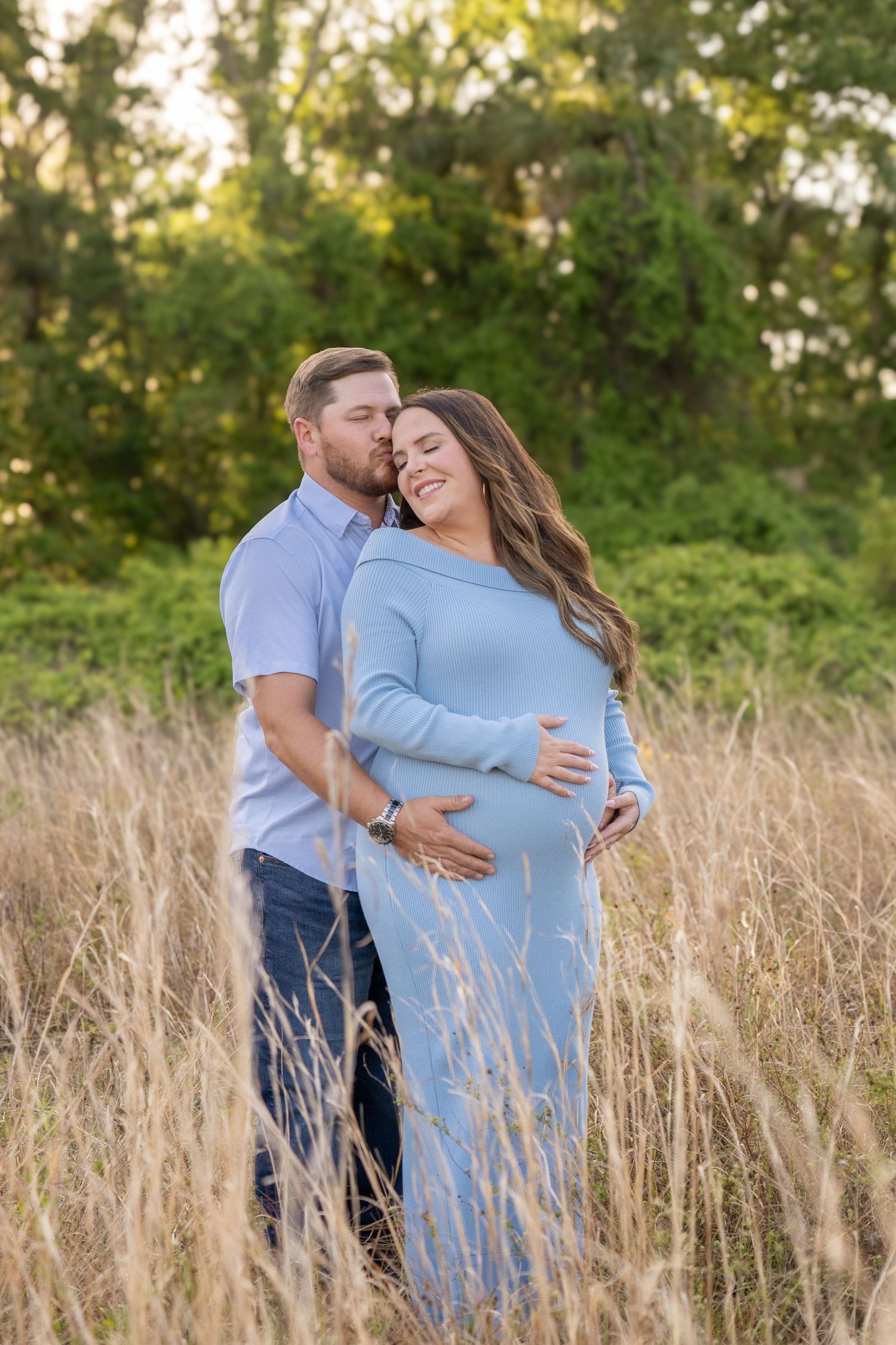 Couple sharing a tender maternity moment in a golden field, woman in blue gown cradling baby bump.